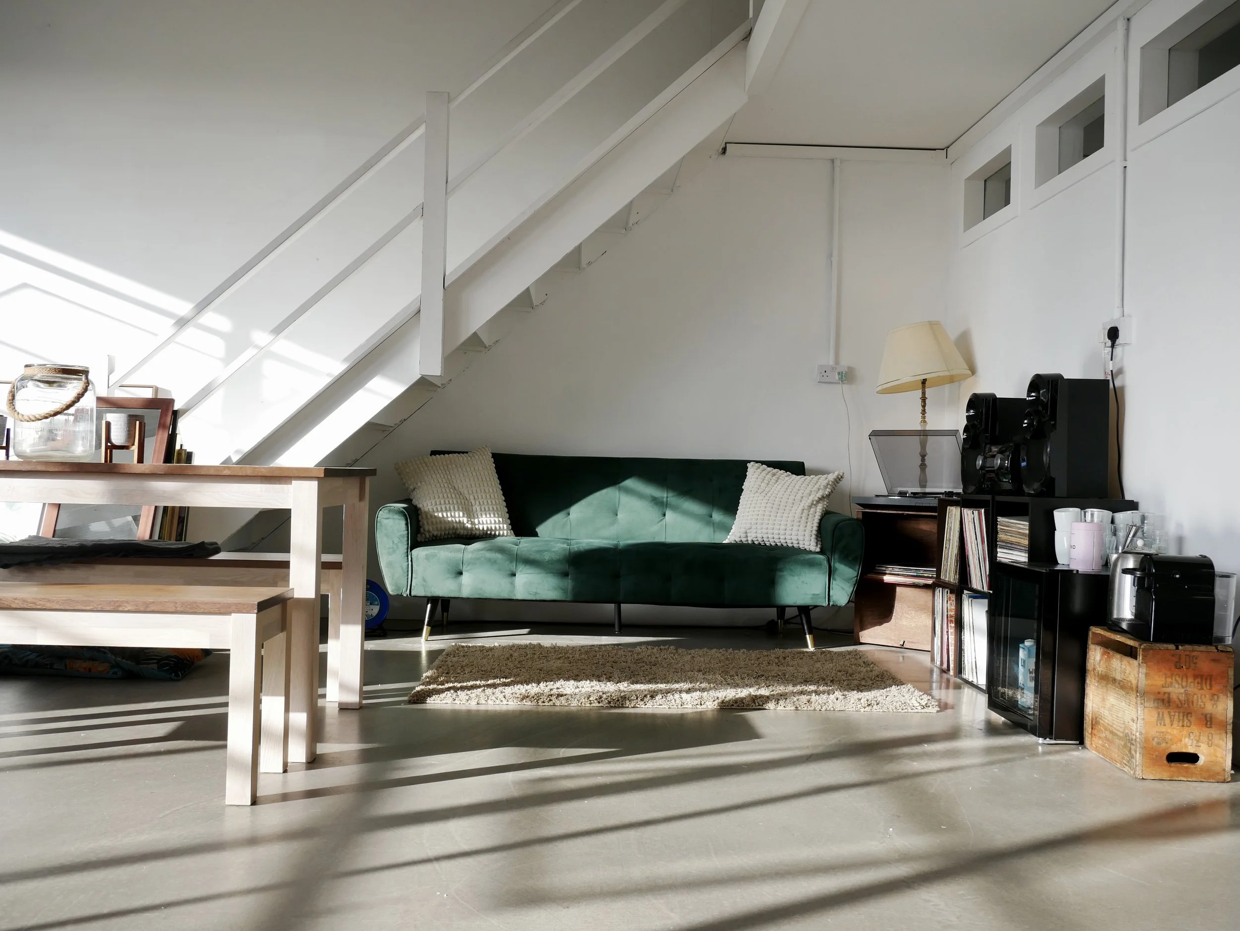 Living room with a green velvet sofa, two beige pillows, a small wooden side table with a lamp and electronic devices, a black bookshelf with vinyl records, a record player, and a box, a beige rug, a wooden dining table, and two chairs near the table
