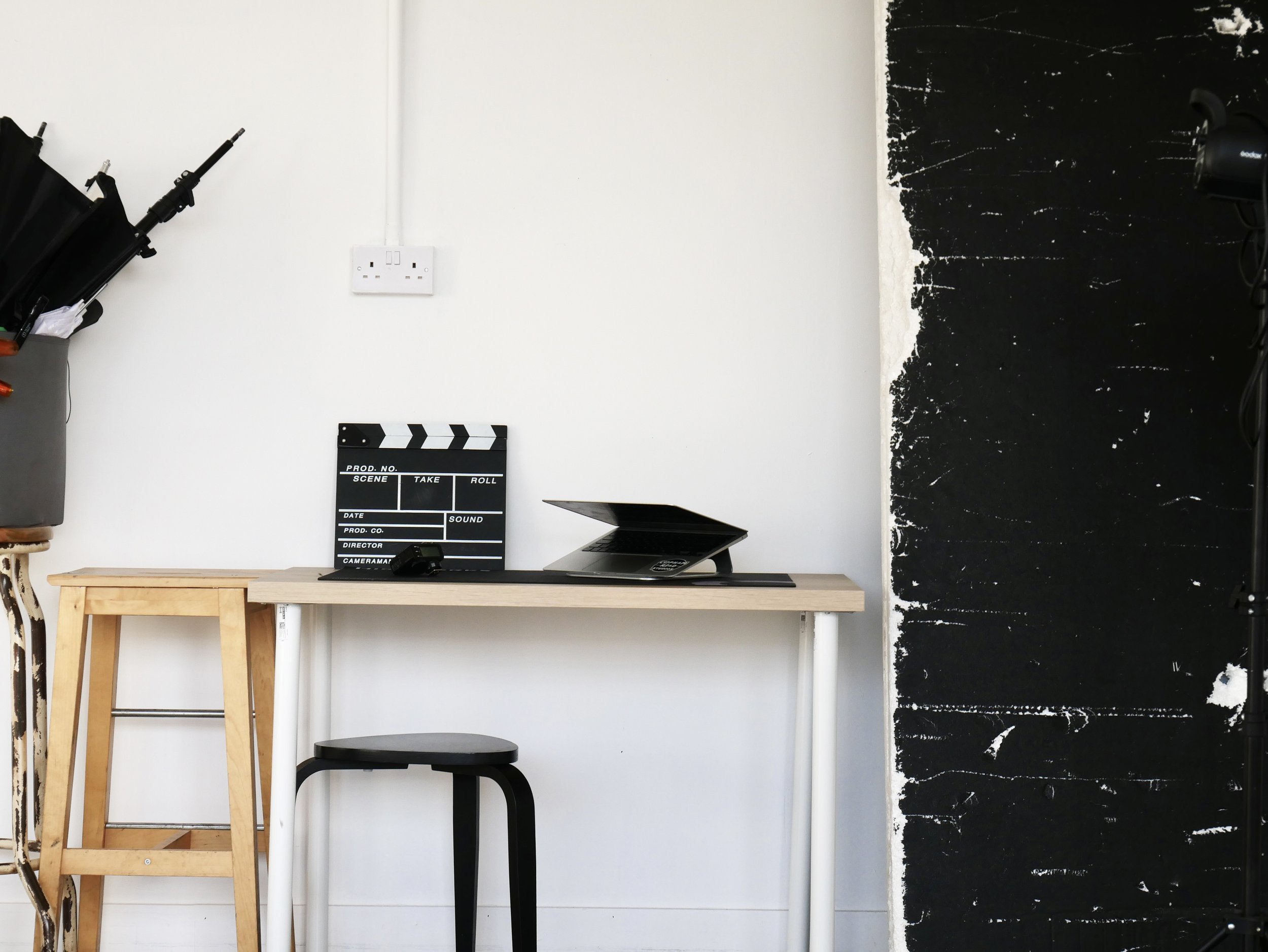 A minimalist workspace with a light wooden desk, a small black stool, a clapperboard, a laptop, and a black chalkboard wall on the right side.