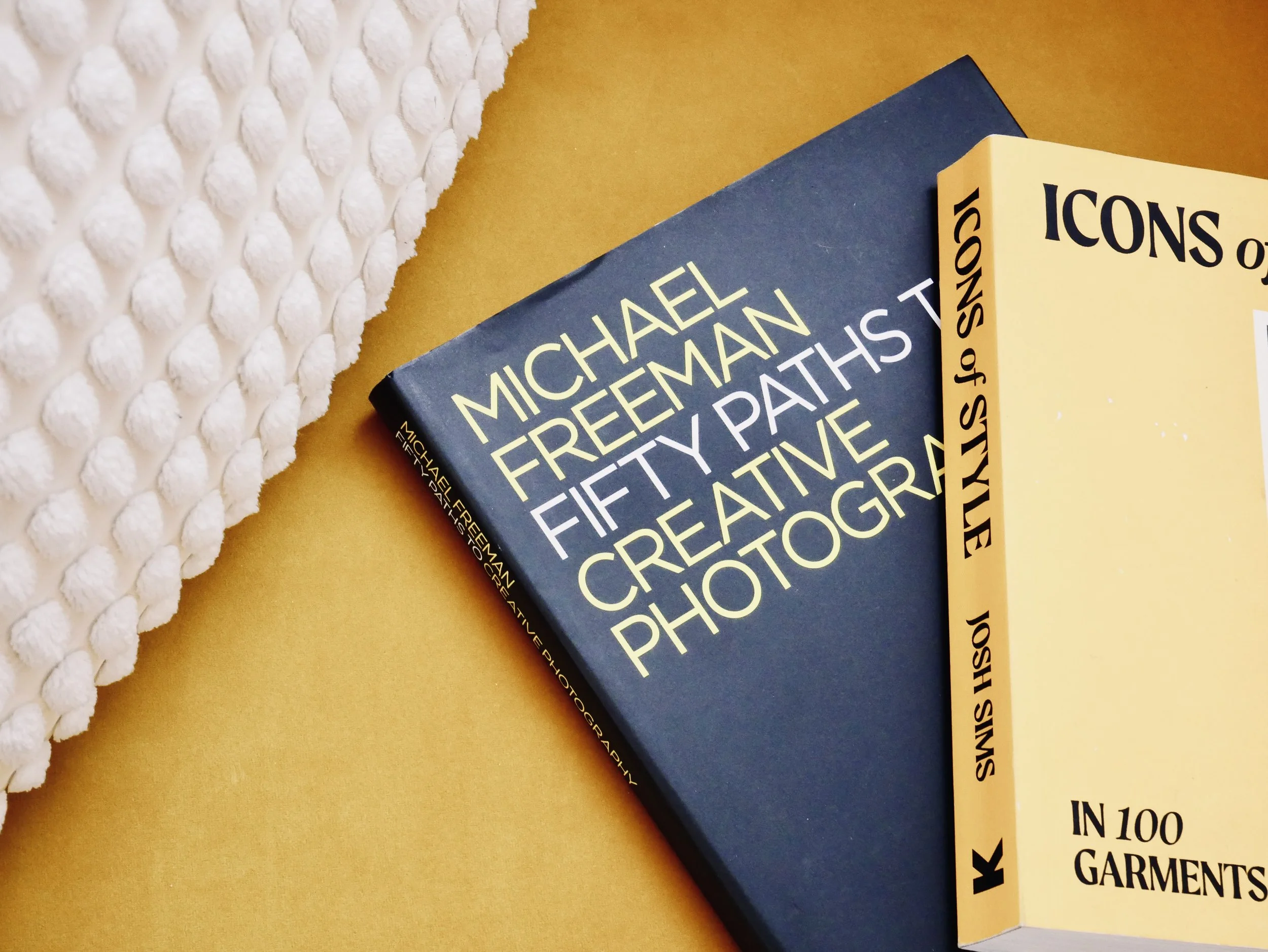 Two books on a yellow surface with a white textured object partially visible on the left. The titles are 'Fifty Path to Creative Photography' by Michael Freeman and another book titled 'Icons of Style in 100 Garments'.
