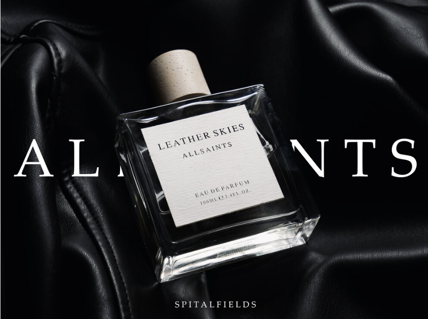 Glass bottle of AllSaints Leather Skies perfume on black leather background with branding text.