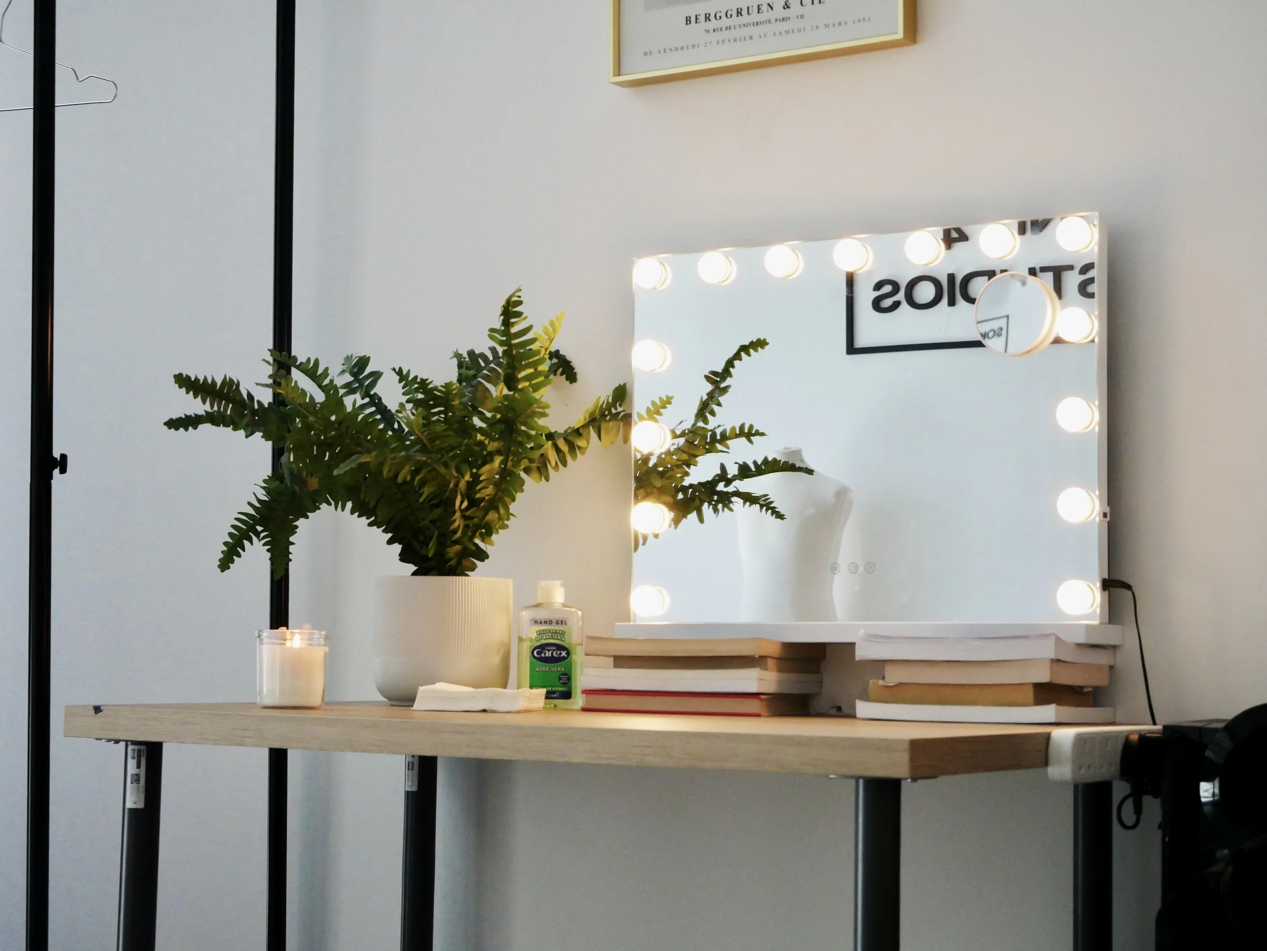 A vanity mirror with bright round bulbs around its border, placed on a wooden table. The mirror reflects part of the room, including a black framed sign and a white mannequin torso. A potted fern, a candle, a bottle of hand gel, and a stack of books are on the table.