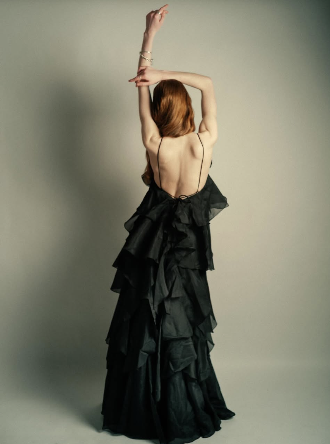 A woman with long red hair wearing a black, flowy, layered dress with thin straps, posing with her back to the camera and arms raised above her head against a plain light-colored background.