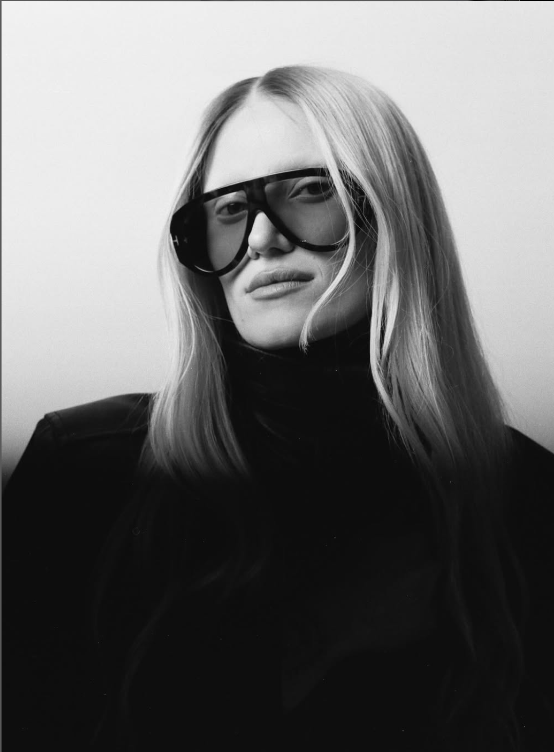 A woman with long, light-colored hair wearing large sunglasses and a black turtleneck, posing against a plain background.