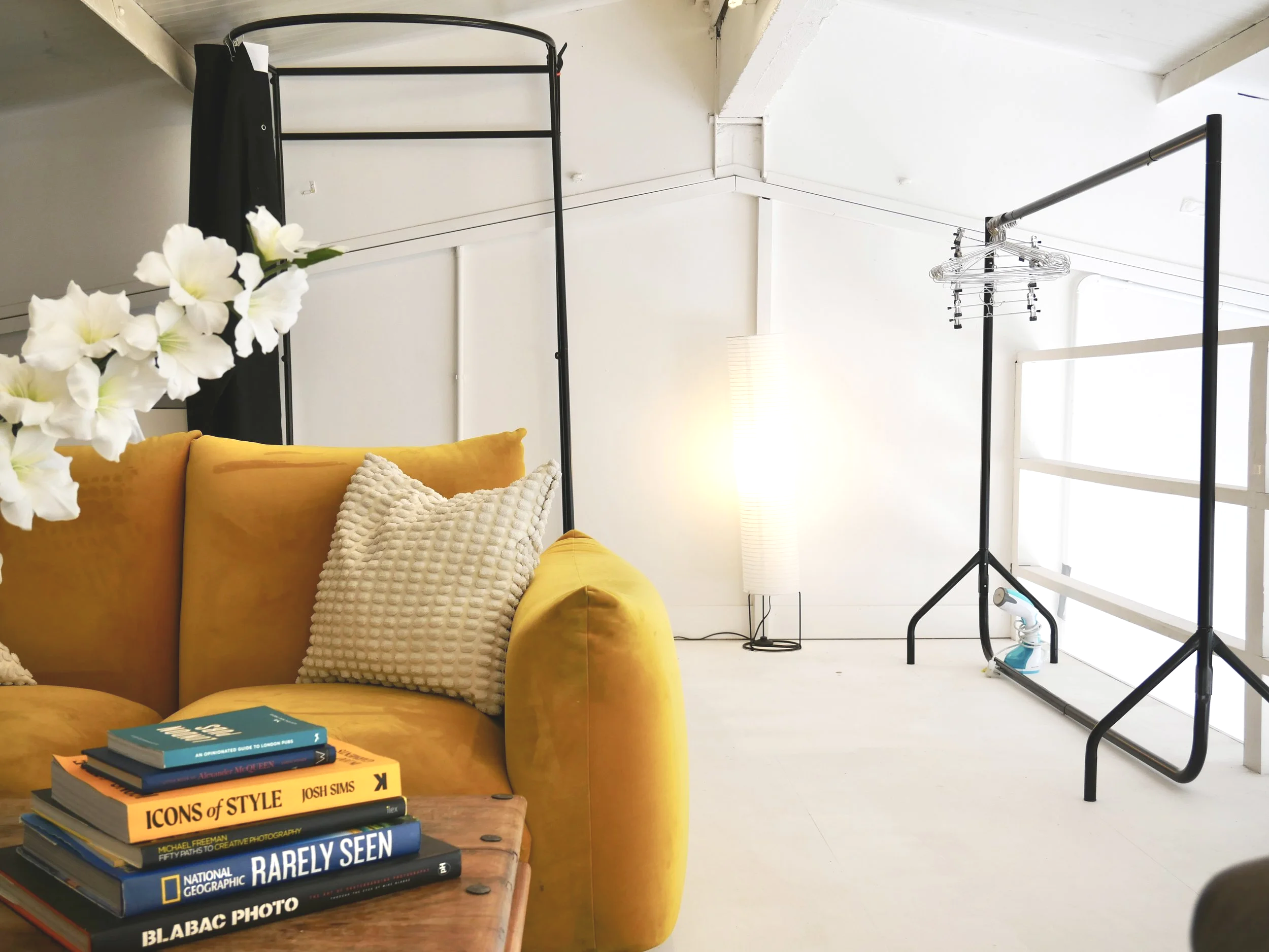 Interior space with a yellow sofa, white pillows, stacked books, and a clothing rack with hangers. A tall white floor lamp and a window providing natural light are also visible.