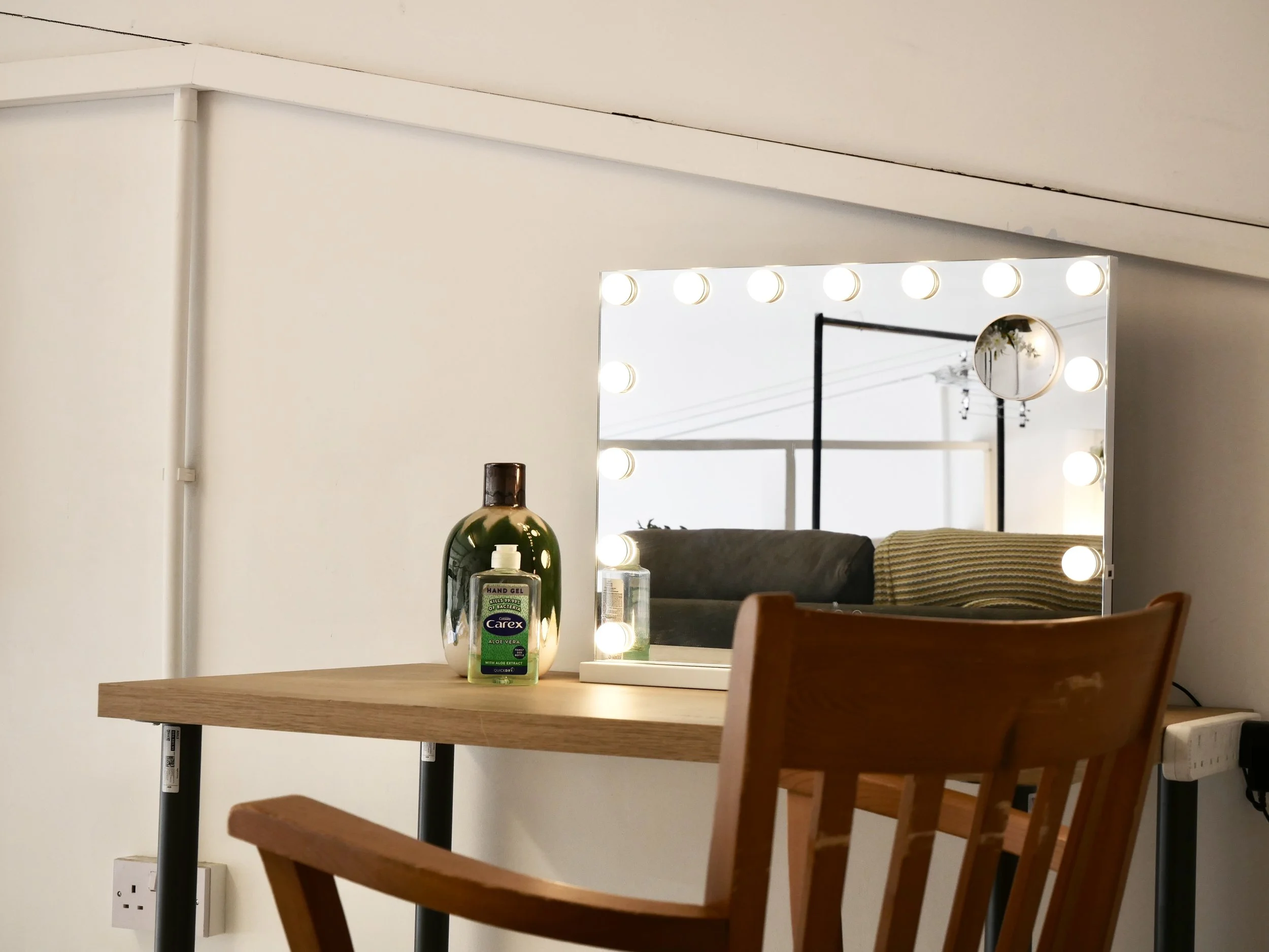 A vanity mirror with lights on a table, with a bottle of hand gel and a lotion bottle in front, and a wooden chair beside it.