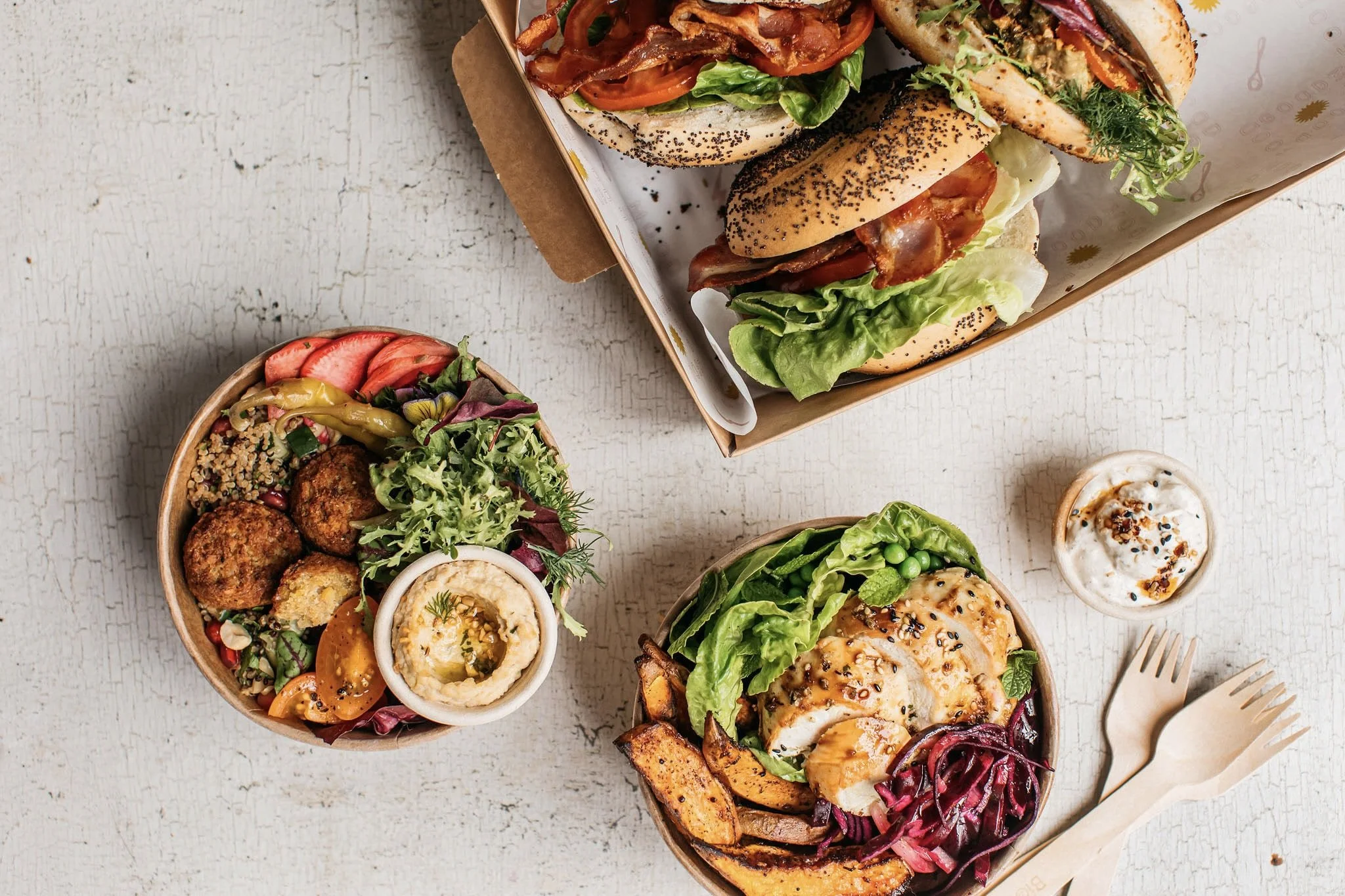 A variety of food dishes on a white textured surface, including a bowl of falafel with salad and hummus, a grilled chicken salad with mixed greens, and a basket of assorted sandwiches with bacon, lettuce, tomato, and other toppings.