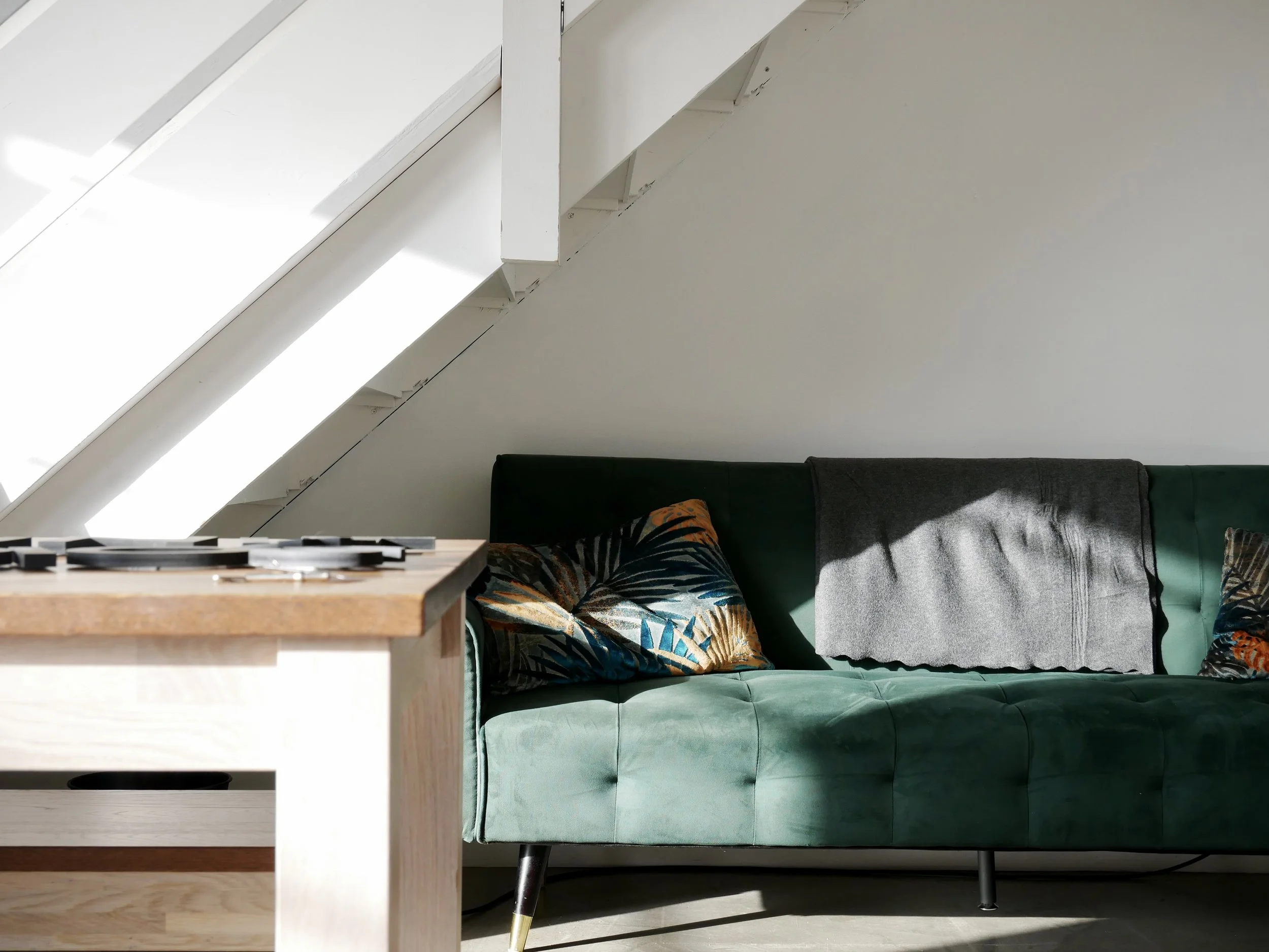 Living room with a green sofa, colorful decorative pillows, a gray blanket, and a wooden table in front, under a sloped ceiling with skylights.