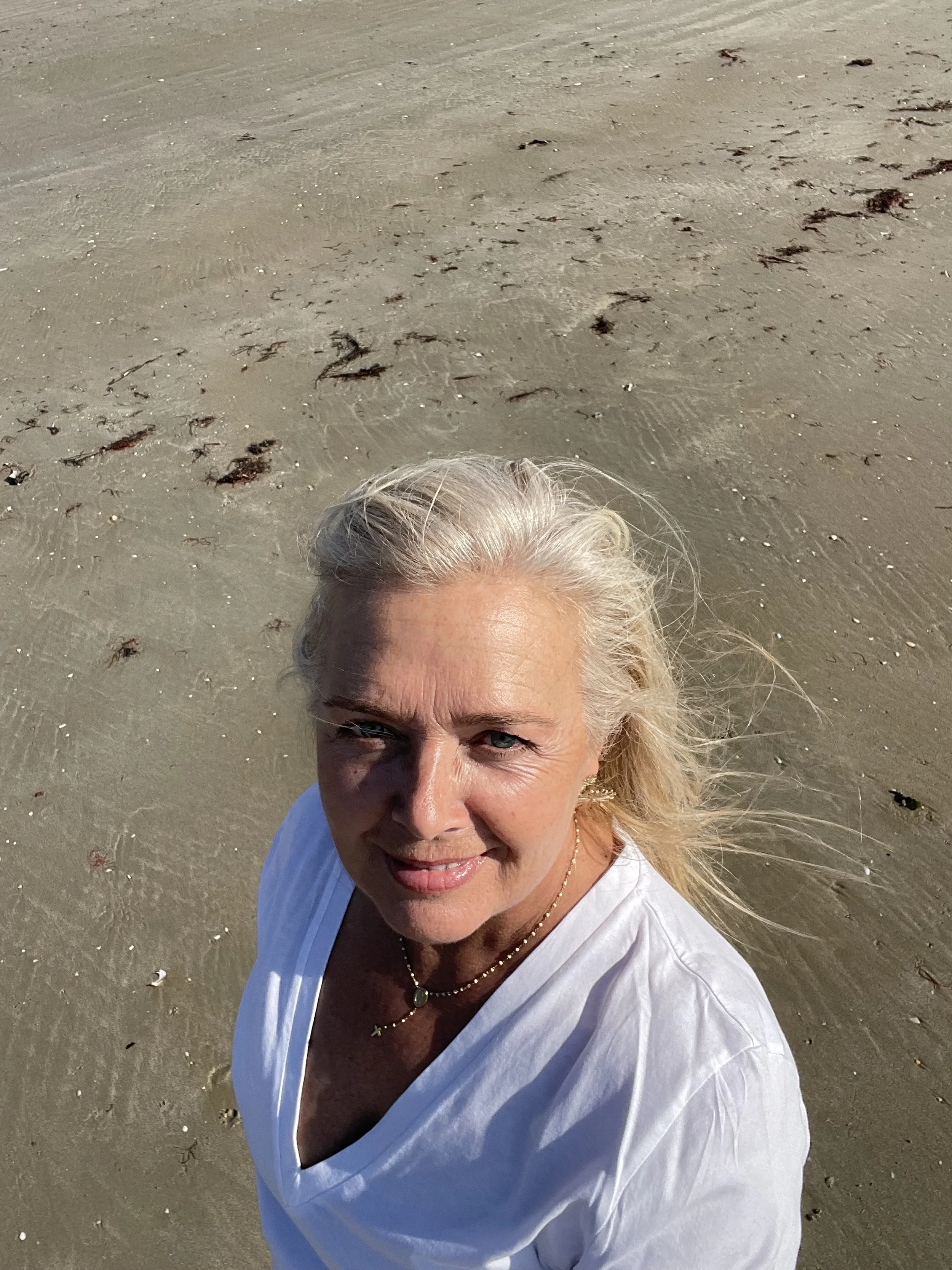 Une femme  fabienne delbouis aux cheveux gris clair sourit sur la plage avec du sable et des algues en arrière-plan. c'est la plage de sainte barbe