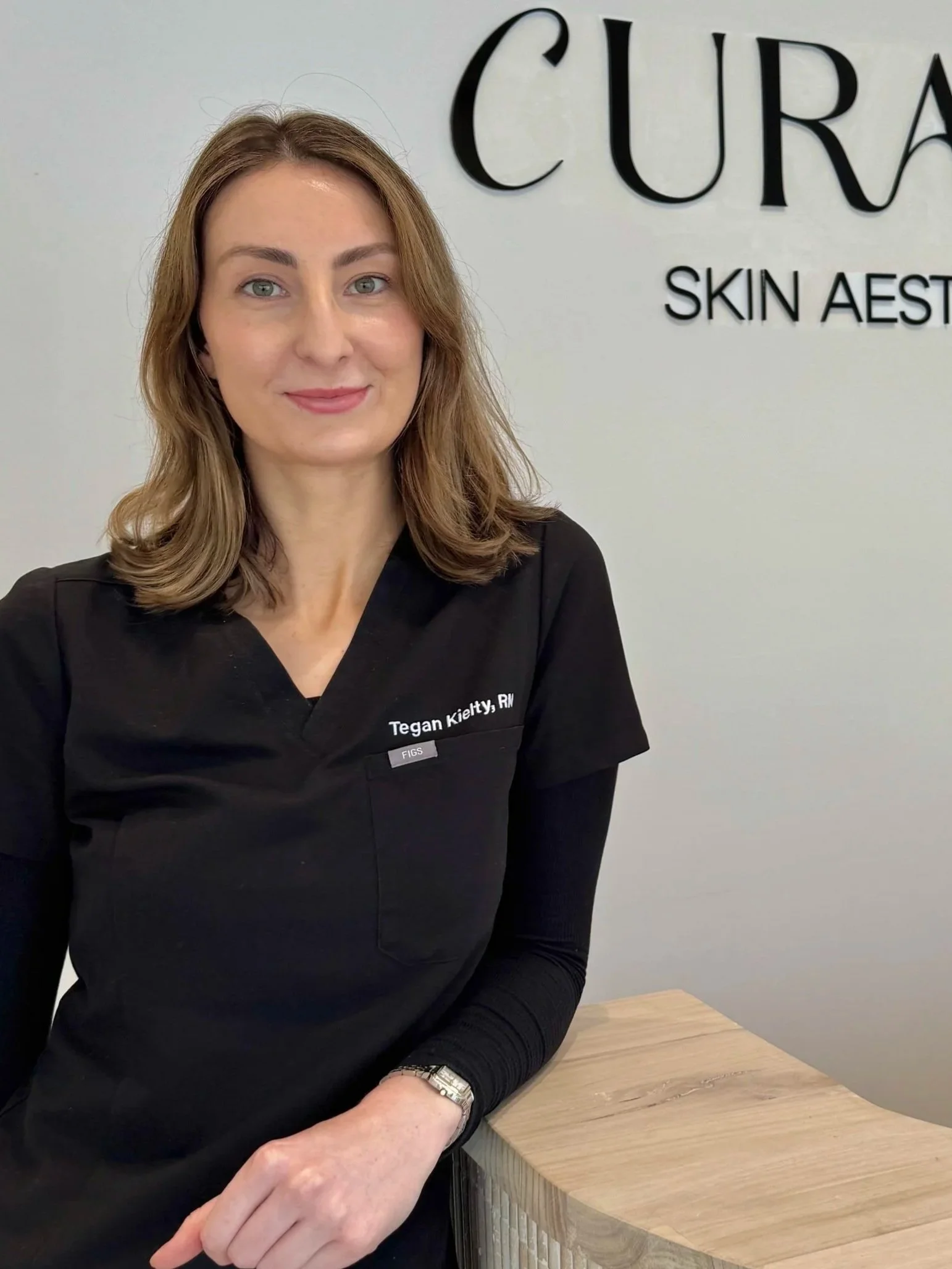 Woman in black scrubs with 'Tegan Kielty, RN' embroidered on it, standing next to a wooden counter in a skin aesthetic clinic with the sign 'CURA SKIN AESTHETICS' on the wall behind her.
