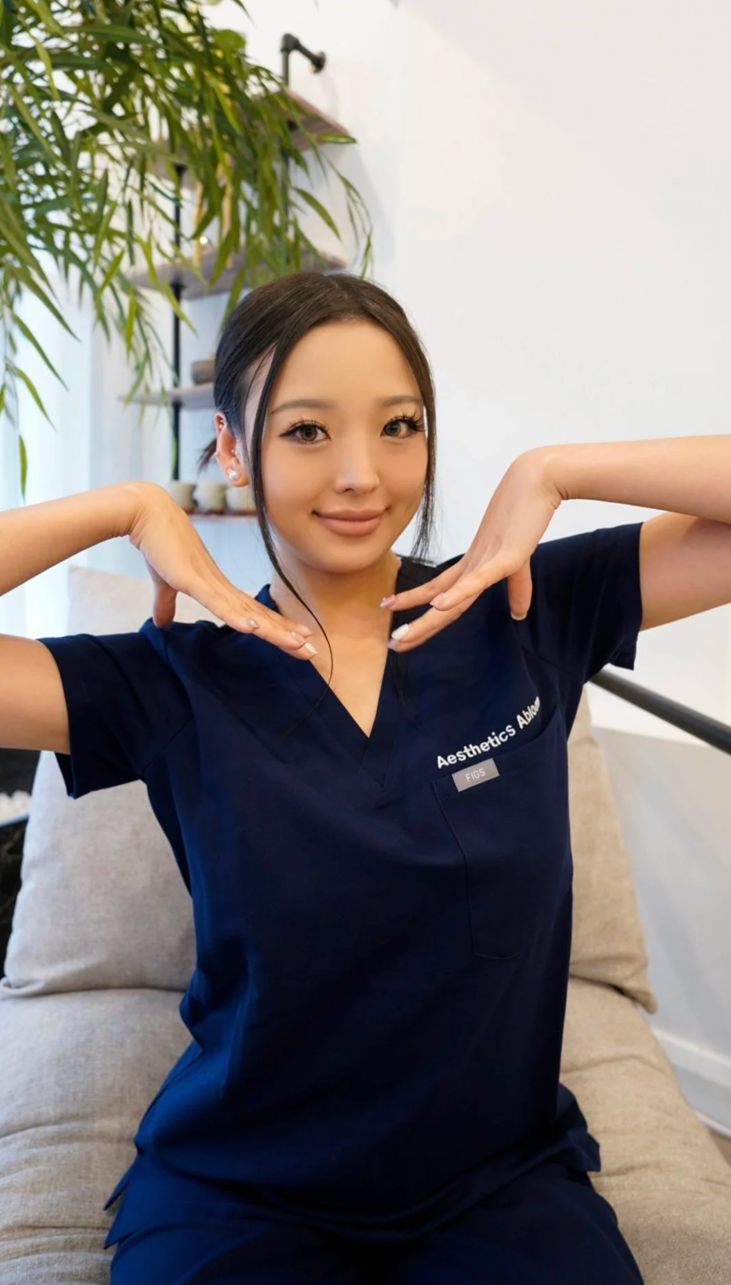 A woman in navy medical scrubs with 'Aesthetics Abia' written on them sits on a beige couch, smiling and posing with her hands under her chin. Behind her, there is a large green plant and a white wall.