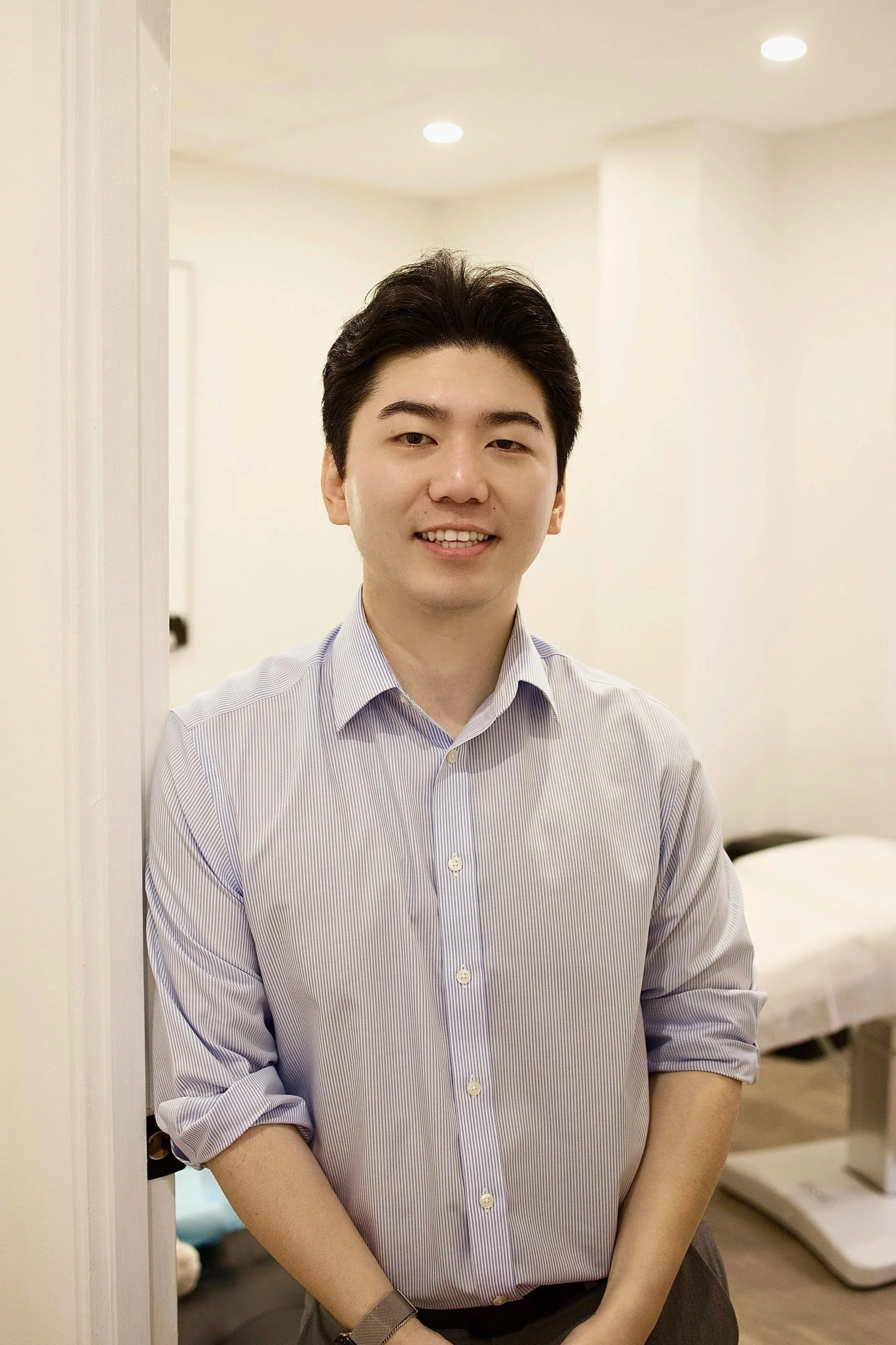 A young man with dark, styled hair smiling and standing indoors, wearing a light purple striped button-up shirt with the sleeves rolled up.