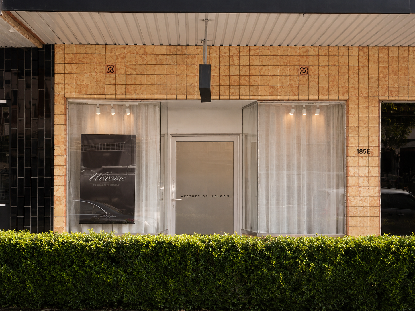 The storefront of a business called Aesthetics Abloom with a glass door and two large windows draped with sheer curtains, a black sign with white text saying "Welcome to your appointment" in one window, and the address 185E on the right wall, with green shrubs in front.