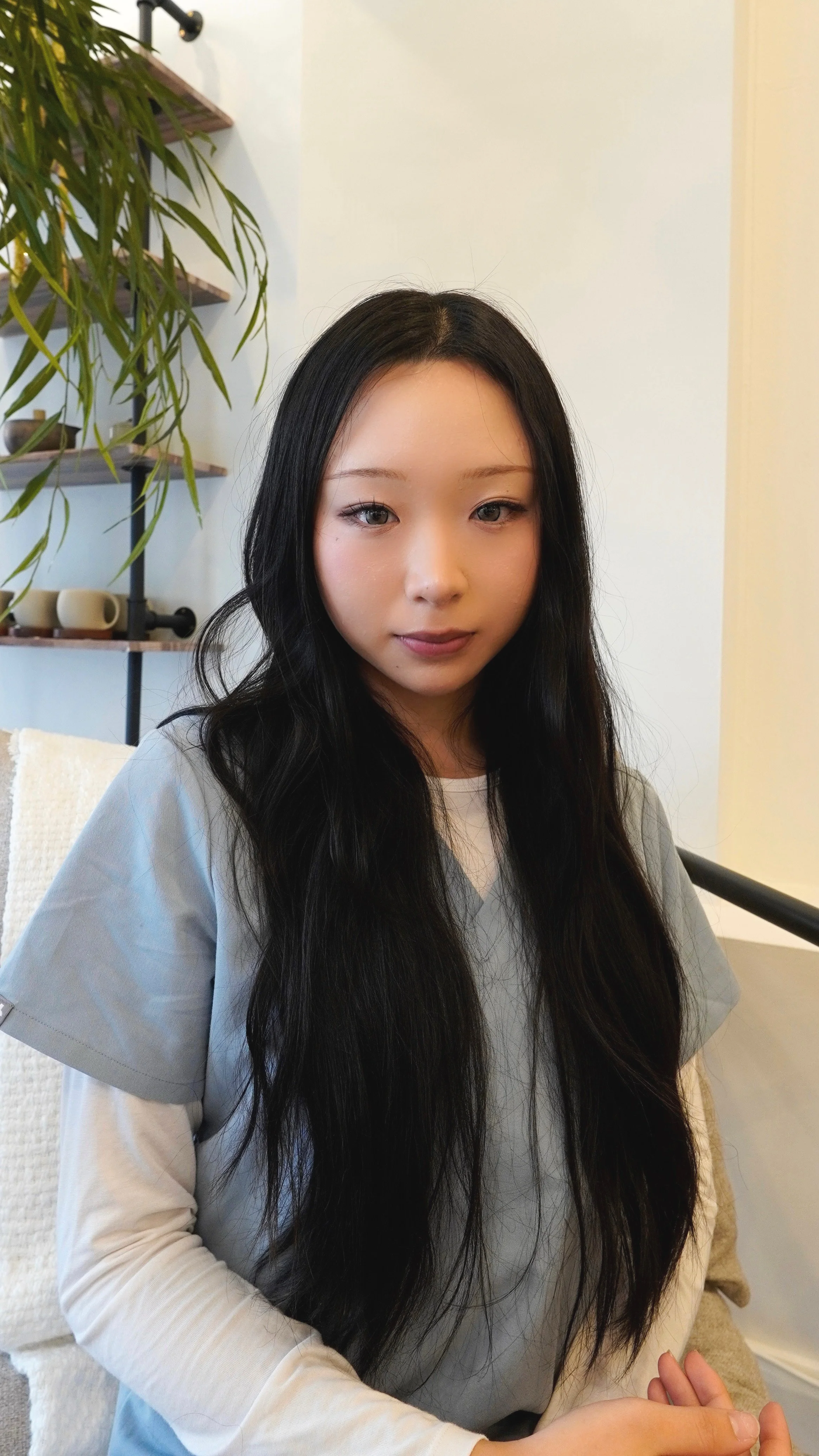 A young woman with long black hair and fair skin sits in a room with a neutral expression, wearing a light gray and white long-sleeve shirt. A plant and shelves with cups are visible in the background.