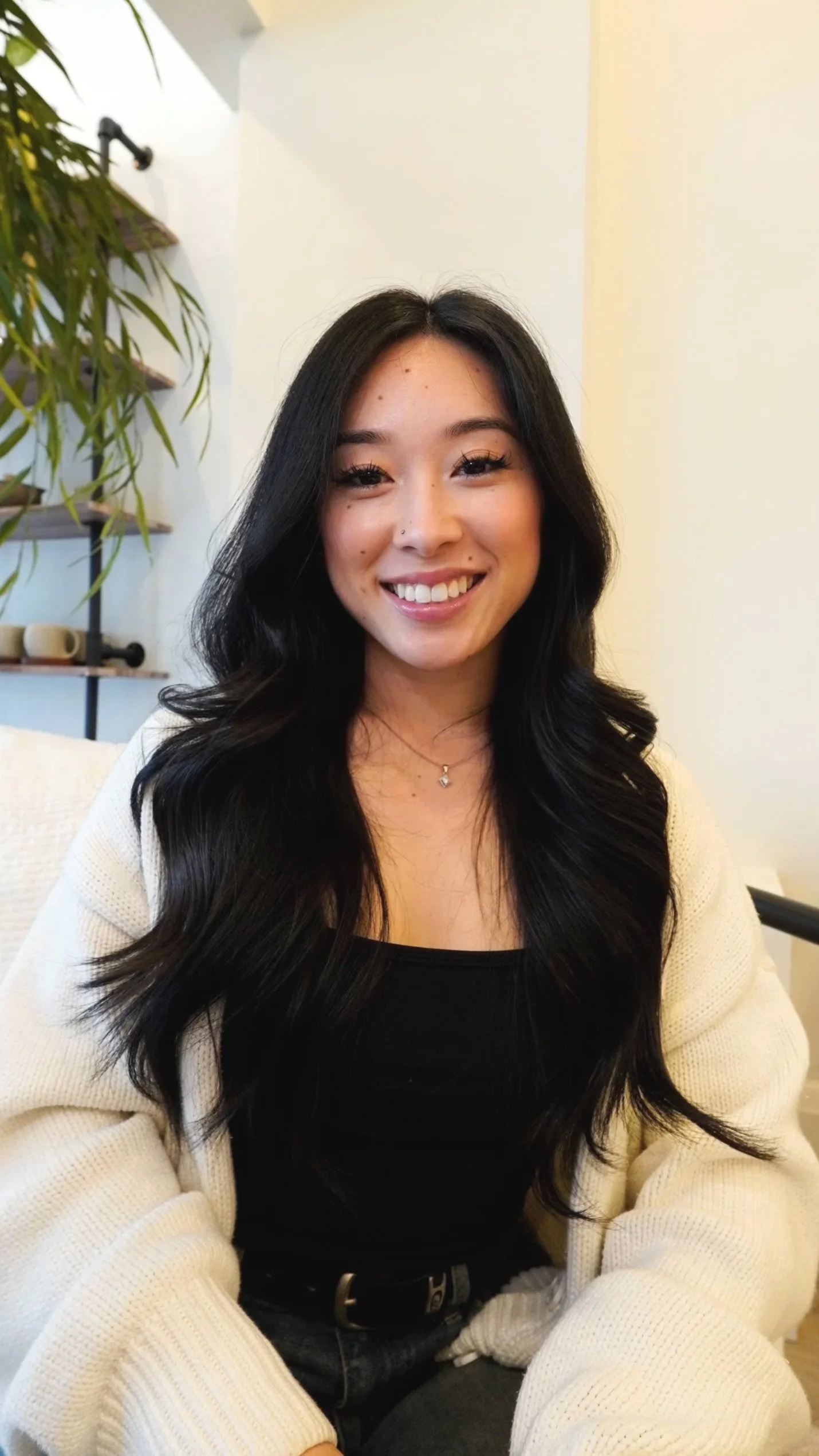A young woman with long black hair, smiling, wearing a black top and a light-colored cardigan, sitting indoors with a plant and shelves with cups in the background.