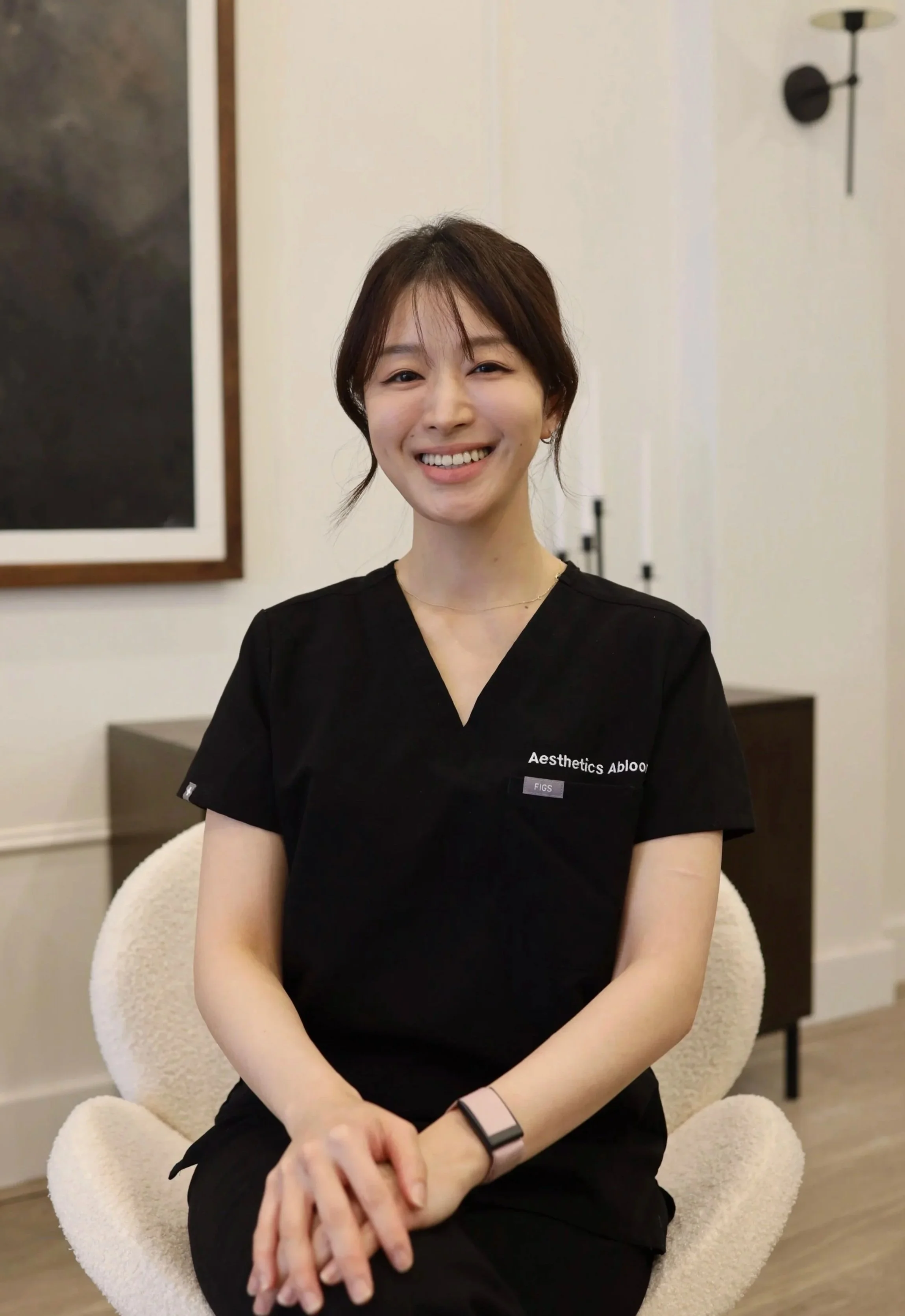 A woman with short brown hair sitting on a cream-colored chair, smiling, wearing a black shirt with white text, in a room with artwork and lighting fixtures.
