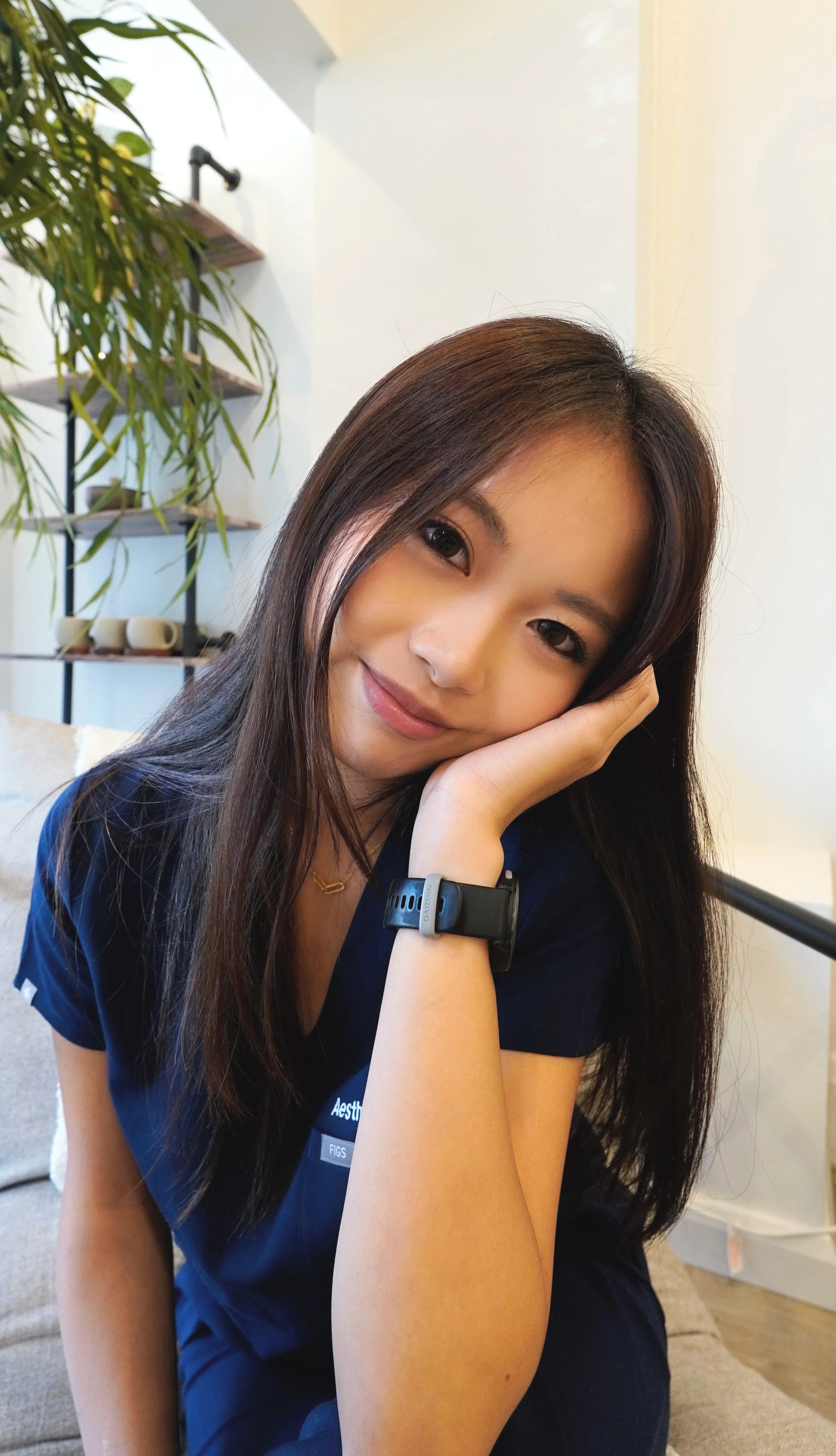 A young woman with long dark hair smiling and resting her head on her hand, sitting indoors near a plant and shelves with cups.
