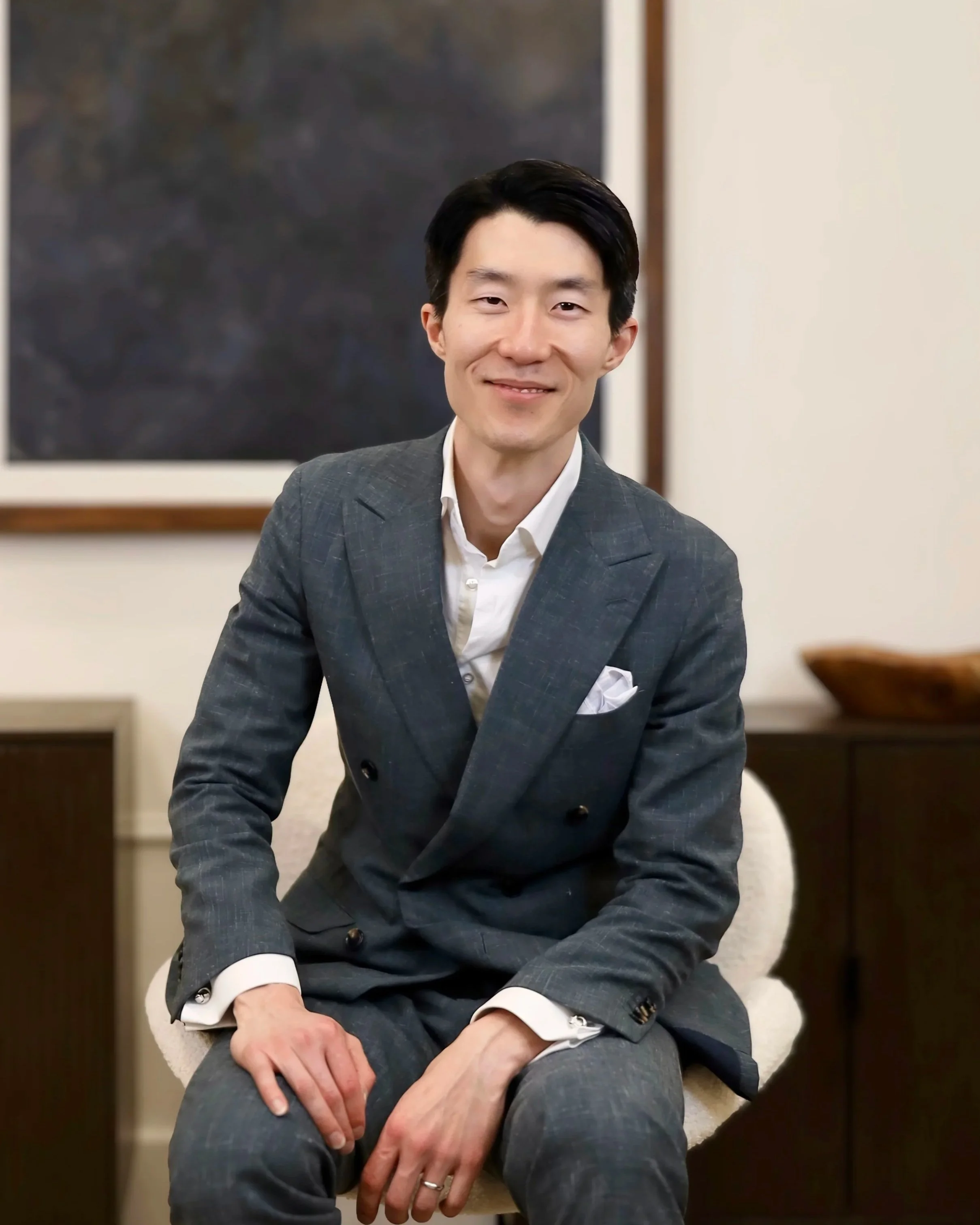 A man wearing a gray plaid suit with a white shirt and cufflinks, sitting on a white chair, smiling at the camera in an indoor setting. There is a dark painting behind him and a wooden piece of furniture to his right.