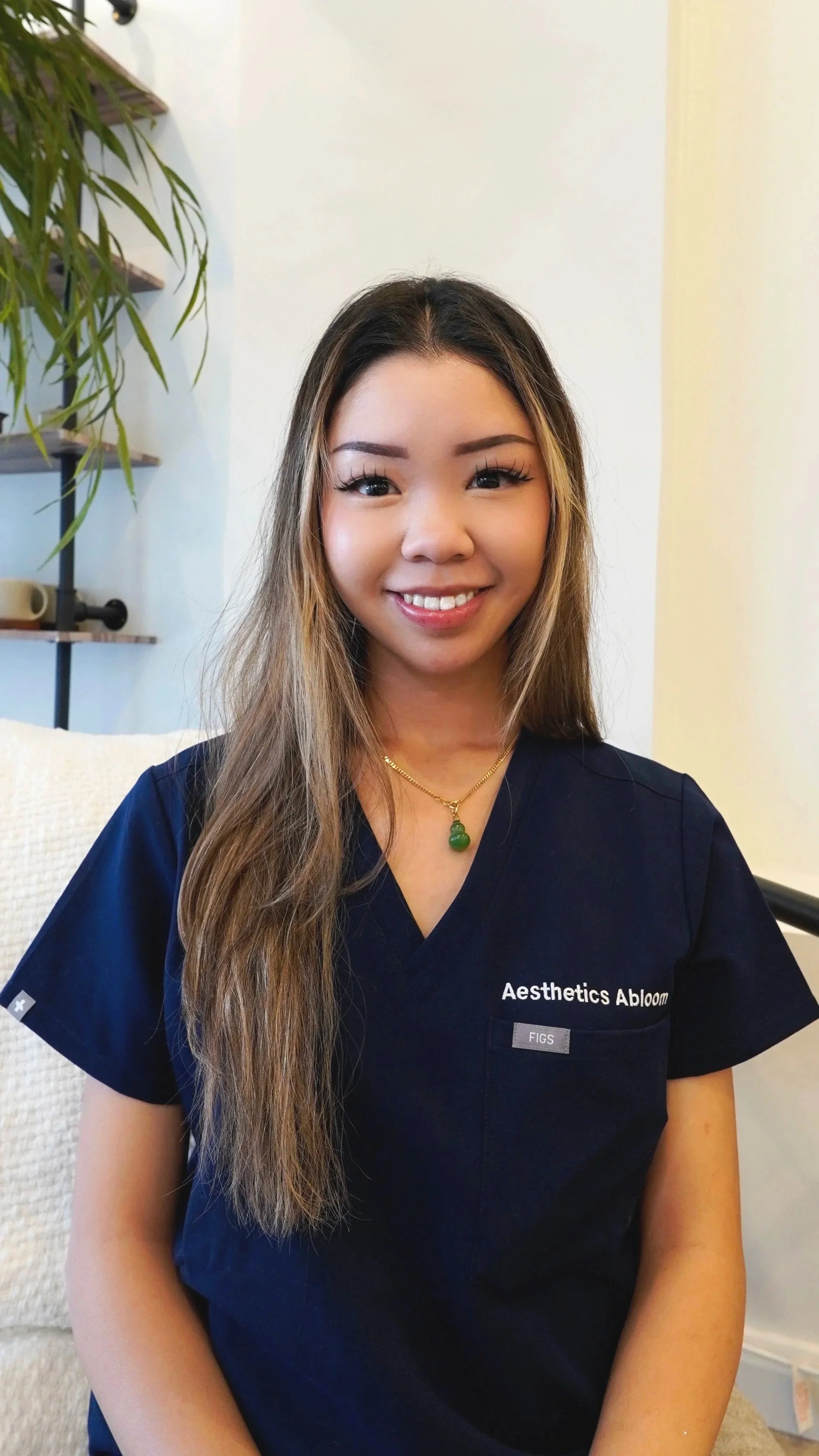 A woman with long, wavy hair wearing a navy medical scrub top with a green pendant necklace, smiling at the camera, sitting indoors with a plant and a white wall in the background.