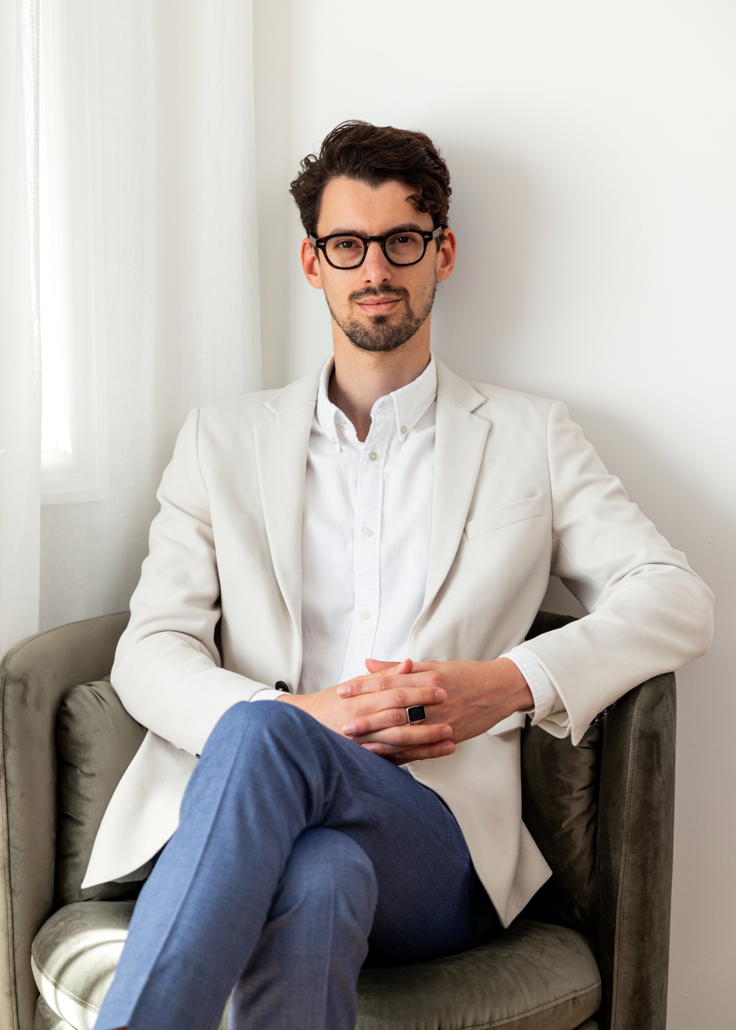 A young man with dark brown hair, glasses, and a beard, wearing a white blazer over a white shirt, sitting on a beige armchair with his legs crossed and hands clasped. He is in a bright room with white walls and a window on the left.