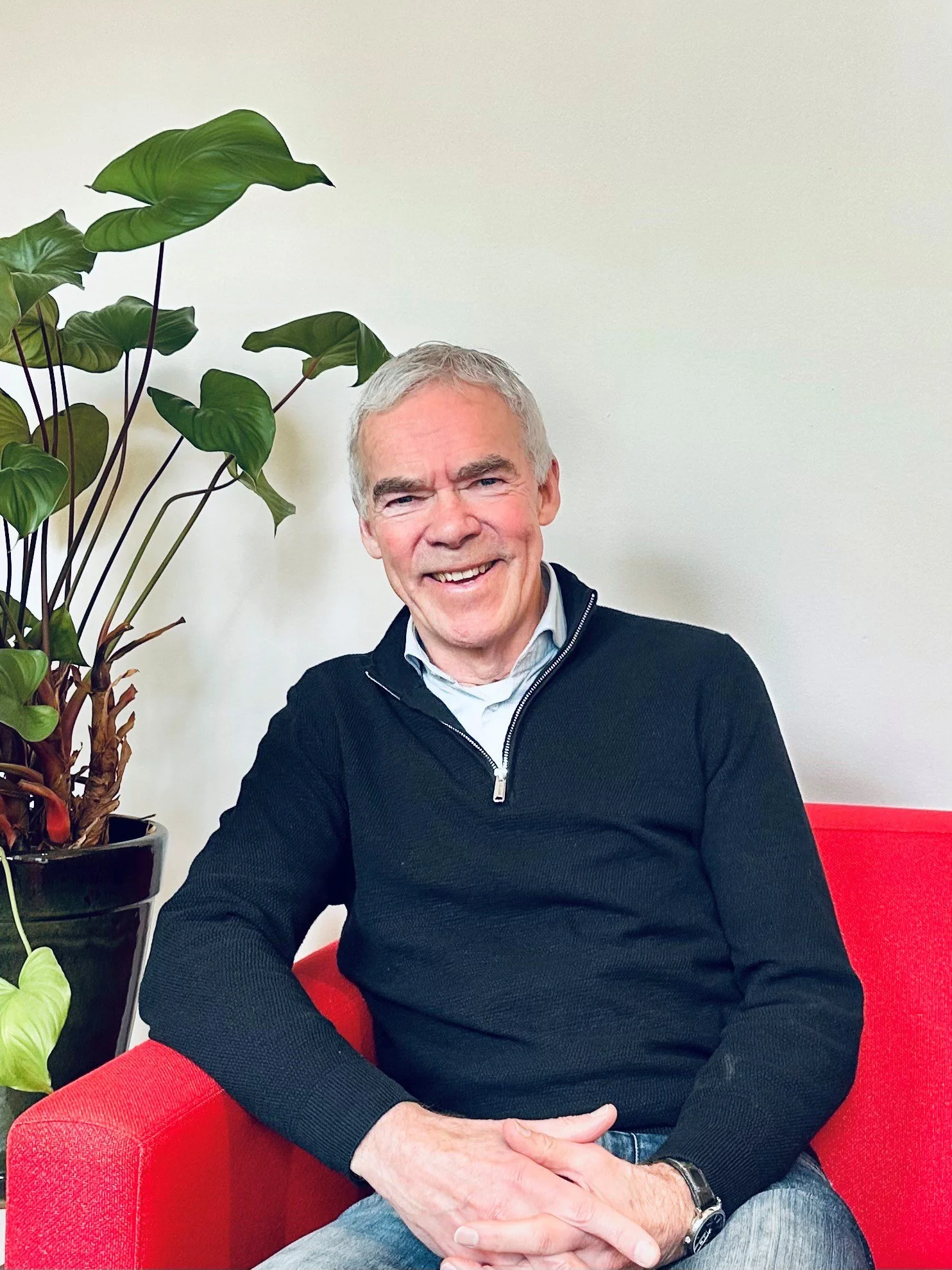 Jan zit op een rode bank in een kamer met een groene plant op de achtergrond. Hij draagt een lichtgrijze polo en heeft een glimlach op zijn gezicht.