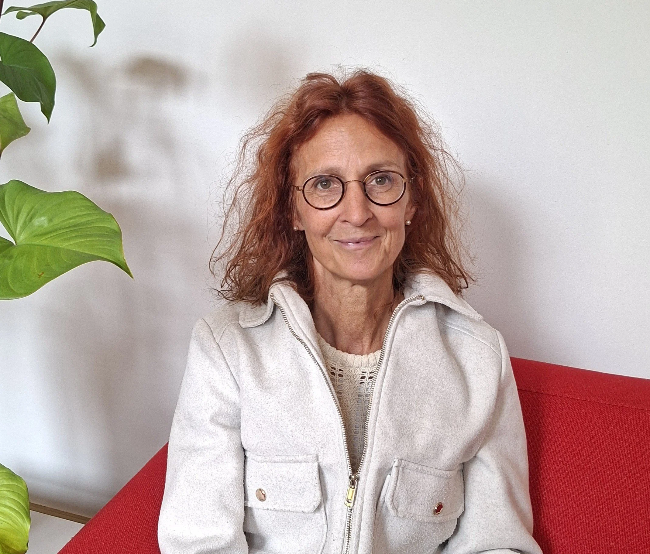 Catelijne, zittend op een rode bank, in een lichte kamer met groene plant op de linkerzijde.