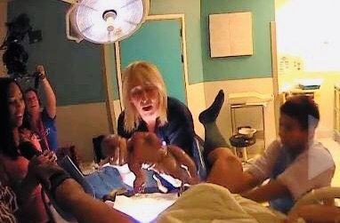 A woman with blonde hair in a blue shirt is surrounded by children, some sitting and some standing, in a brightly lit room with green walls and yellow accents. The woman appears to be interacting with the children, possibly during a playful or educat