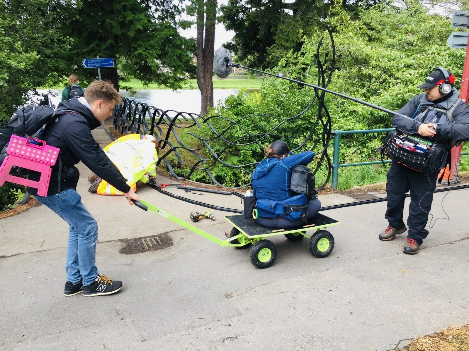 Filmmaking crew setting up camera equipment on a dolly, with one person pulling the dolly while a woman sits on it, and a man operating a boom microphone nearby, in a park with trees and a river in the background.