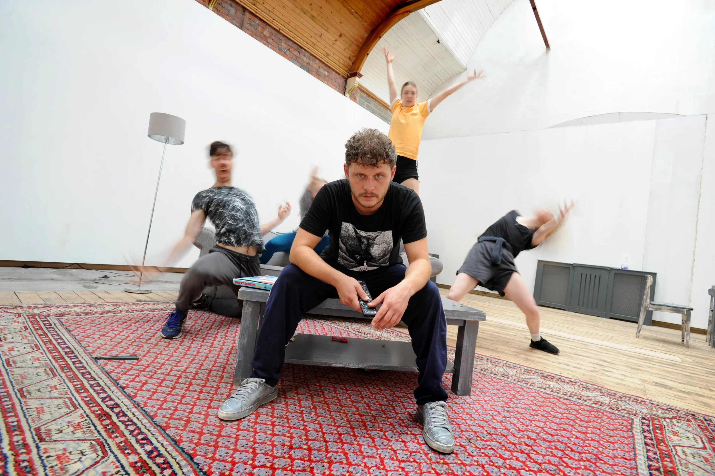 white man is sat on a table set on a red rug staring down the camera. he is surrounded by 3 other dancers reaching out