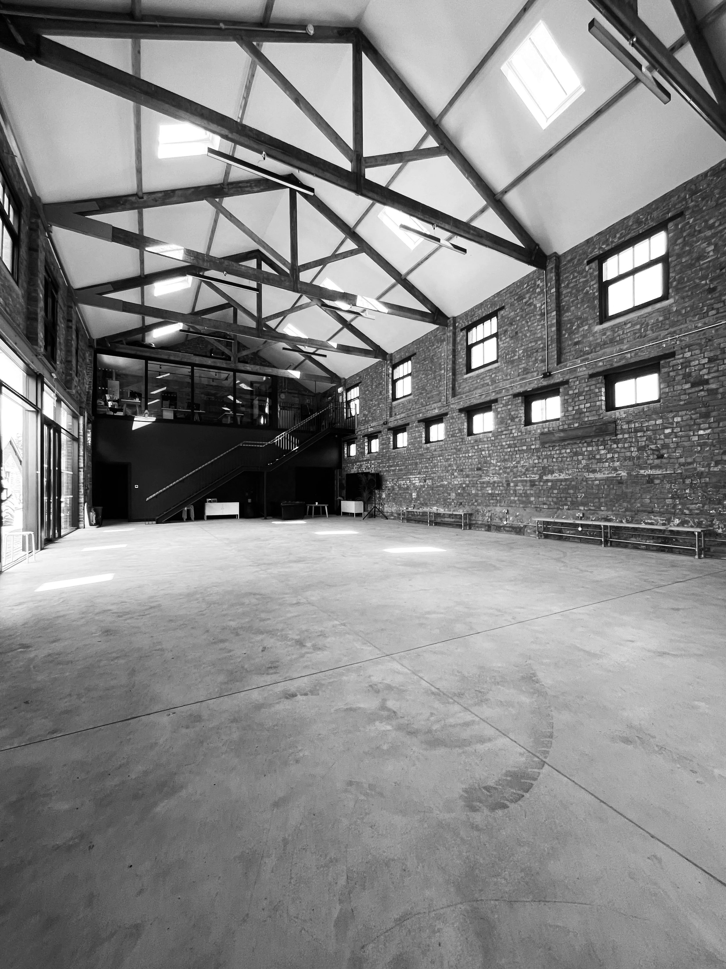 large open studio space with concrete floor, red brick walls and wooden high beams. the second floor has a glass fronted office space