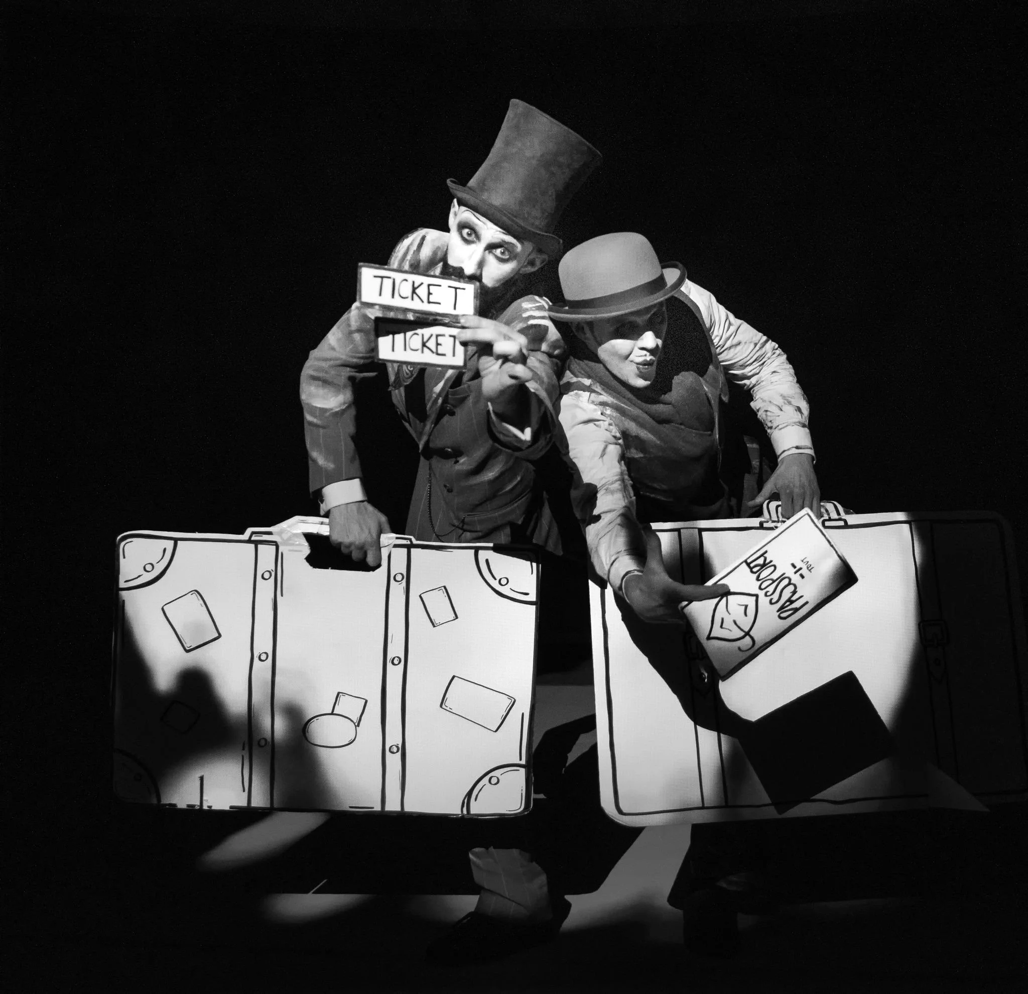 Black and white image of 2 men dressed in suits holding cardboard props of suitcases and tickets