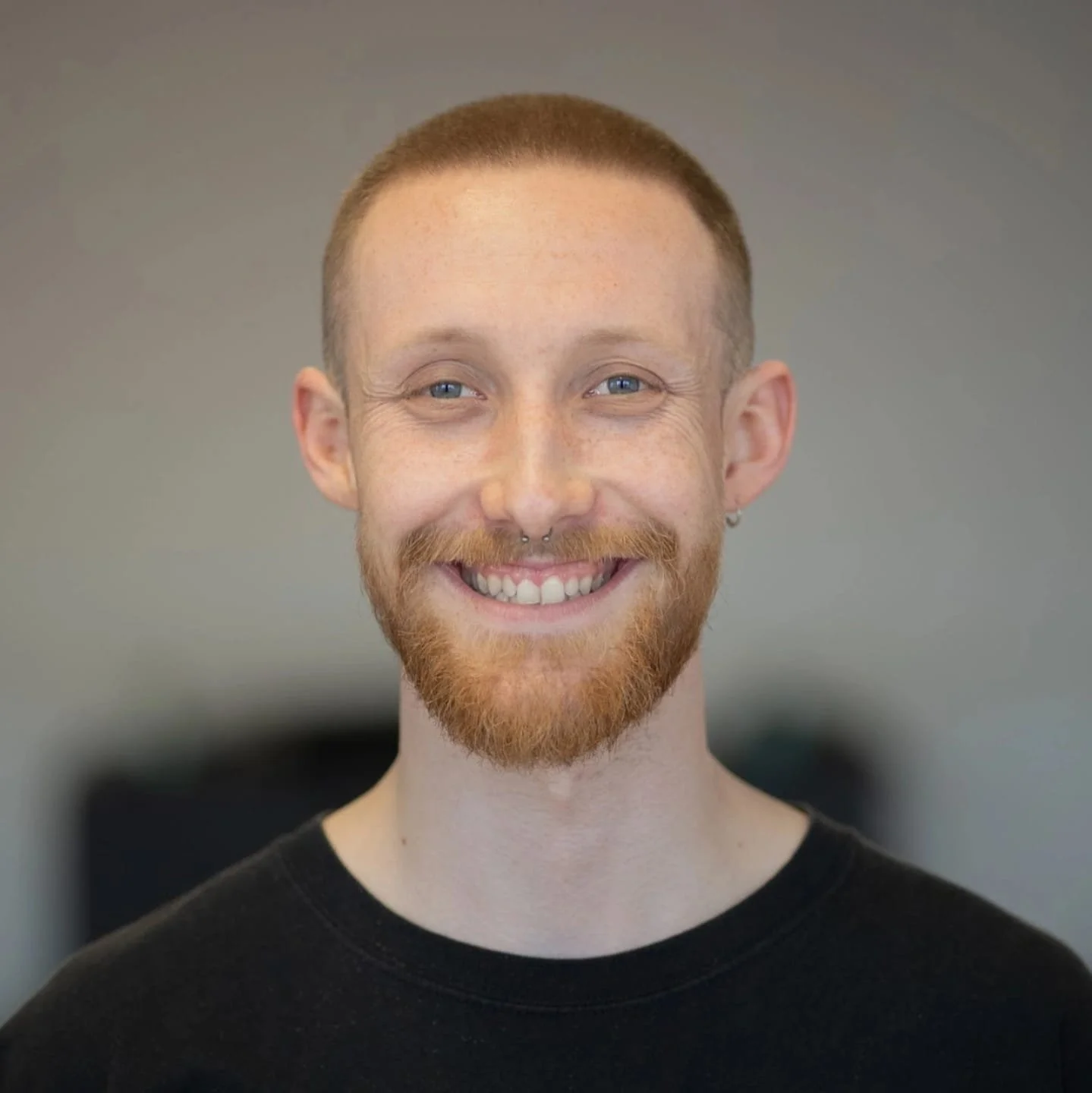 A smiling man with red hair and a beard standing outdoors with a blurred green background.