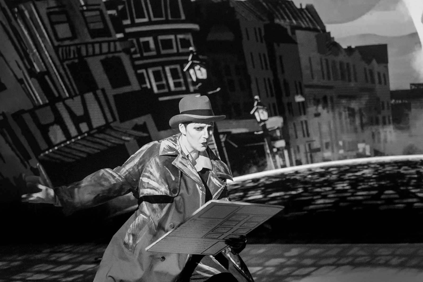 Black and white image of woman in trench coat holding a cardboard prop of a large newspaper. behind is projection of an old street