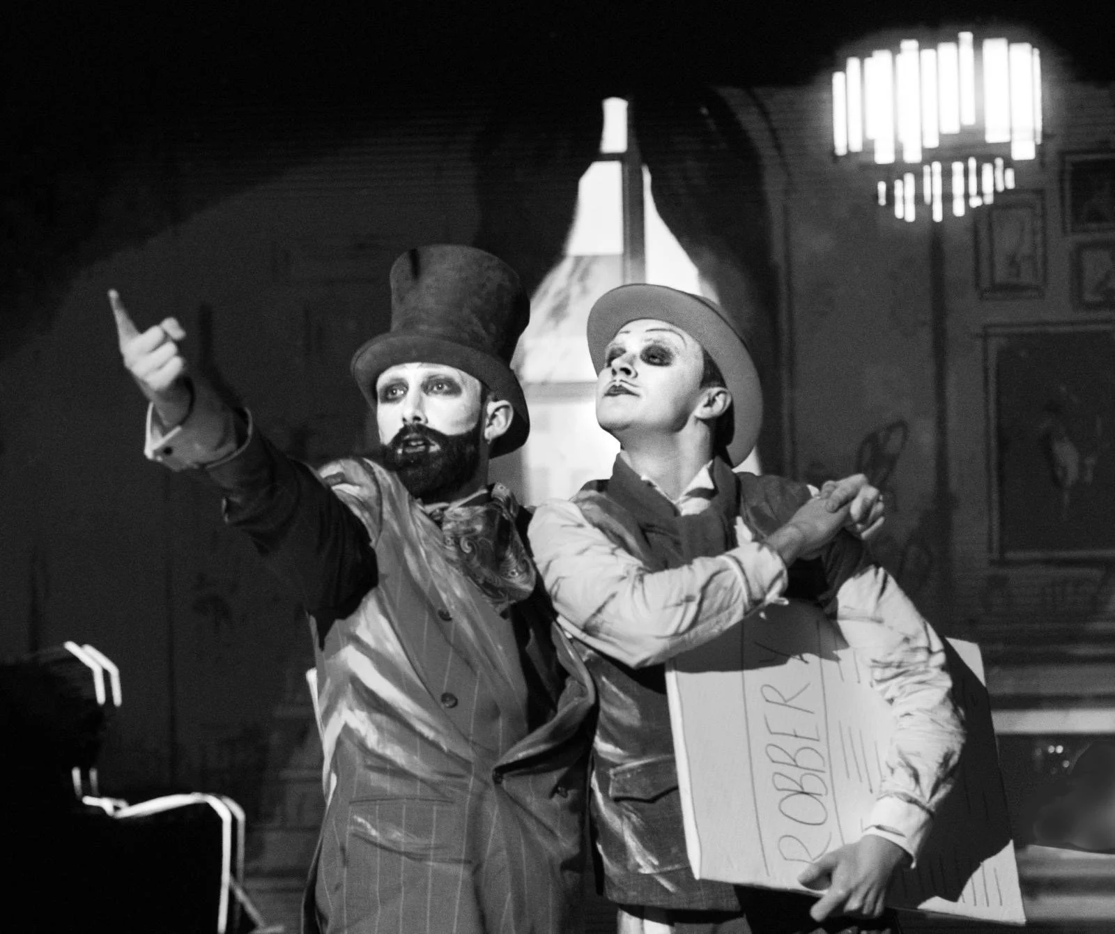 Black and white image of men dressed in suits standing in front of a window and chandelier. One man points at something