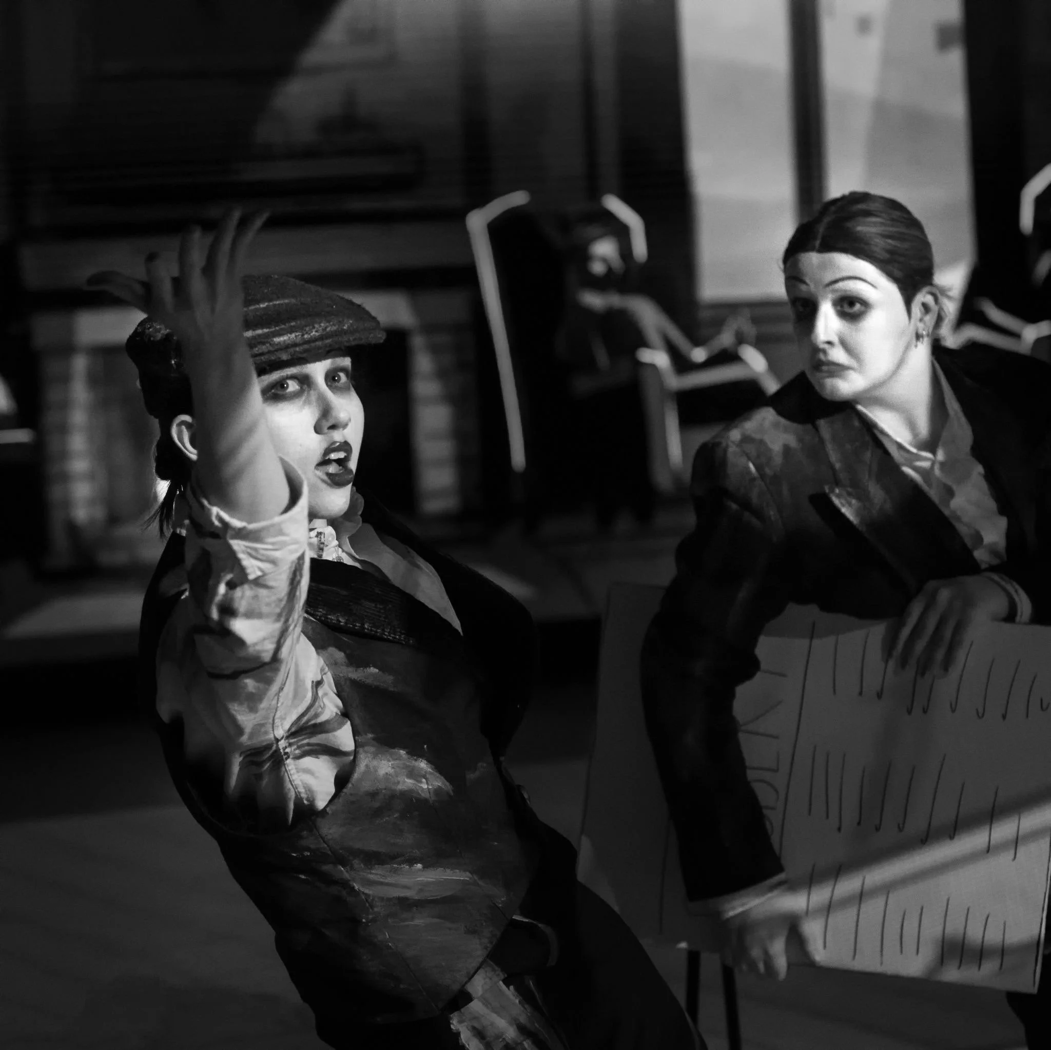 2 women dressed in suits looking surprised. one has arms outstretched the other holds a newspaper