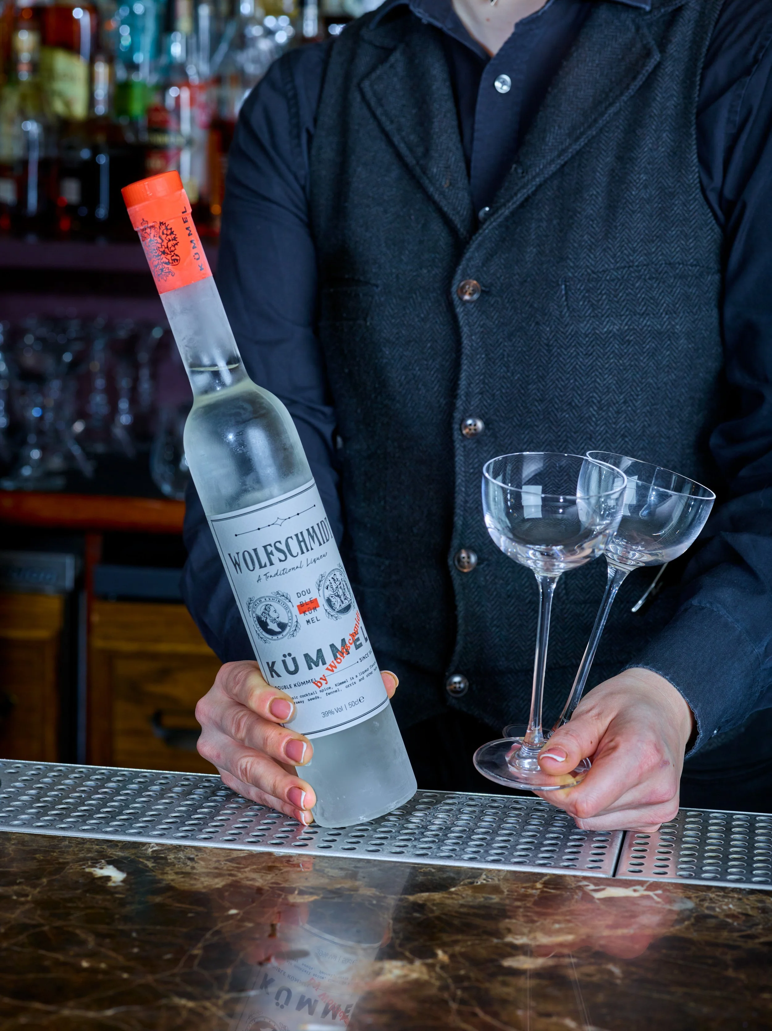 Person holding a Wolfschmidt Kummel bottle with champagne coupe glasses, at a bar