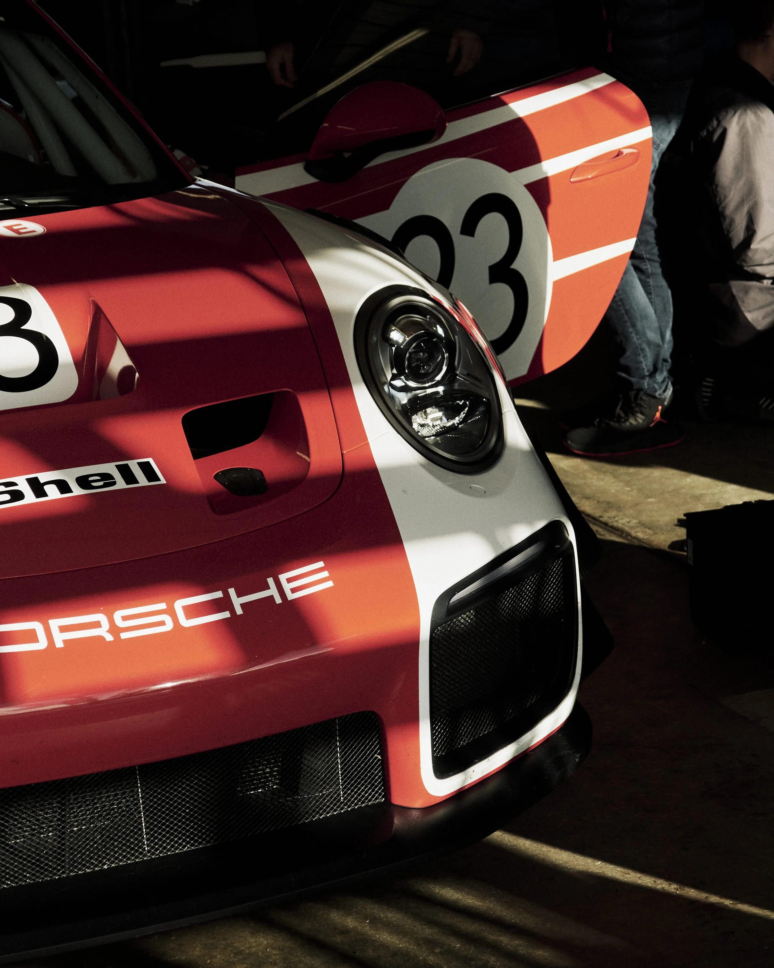 Close-up of a vintage Porsche race car with red, white, and black livery, featuring the number 23 and sponsorship logos, inside a garage or pit area.