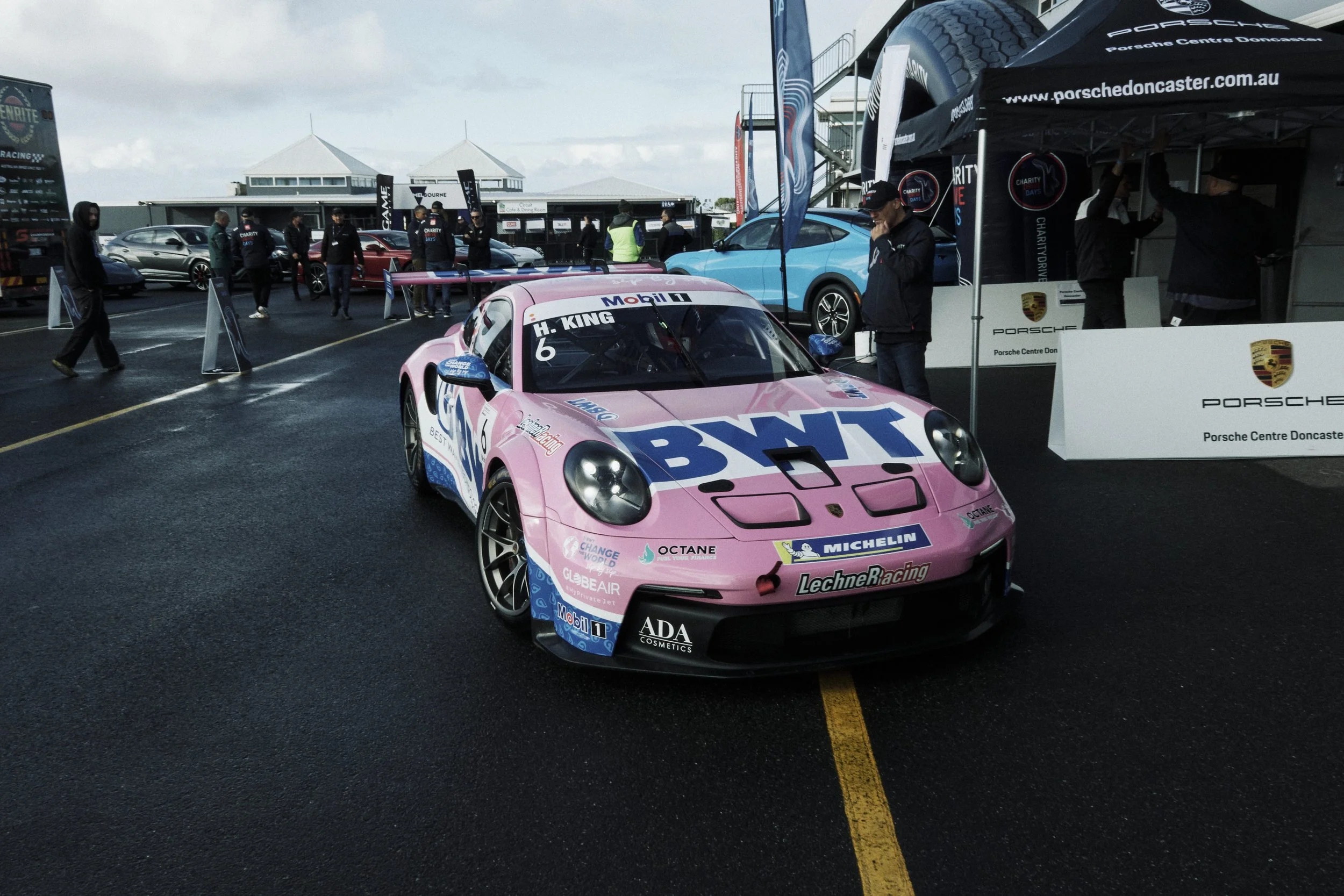 Pink race car with BWT logo and sponsorship decals on a wet race track, surrounded by people and support tents at a racing event.