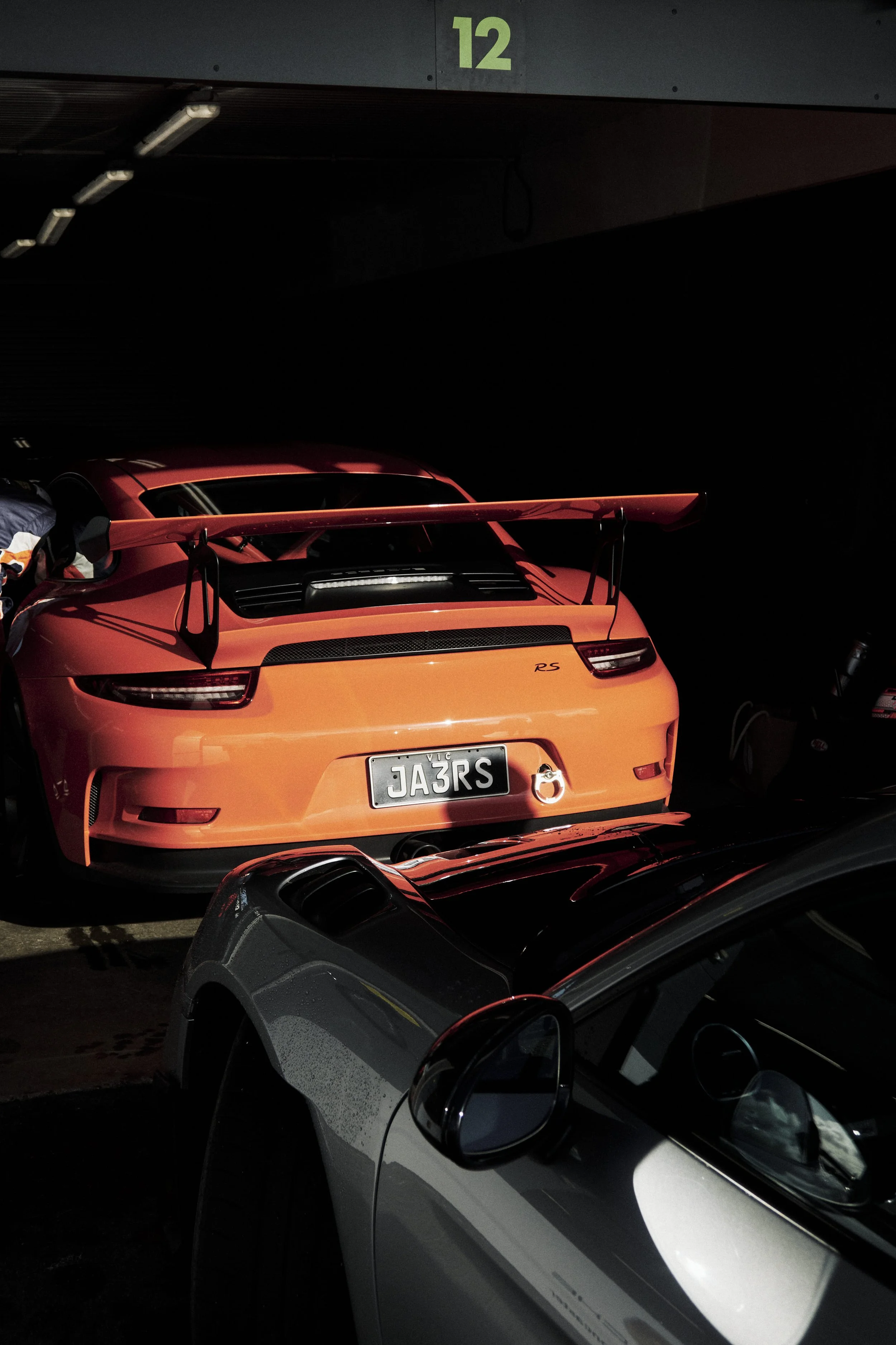 The rear of a bright orange sports car, likely a Porsche 911 RS, parked in a dark parking garage with a green number 12 painted above.