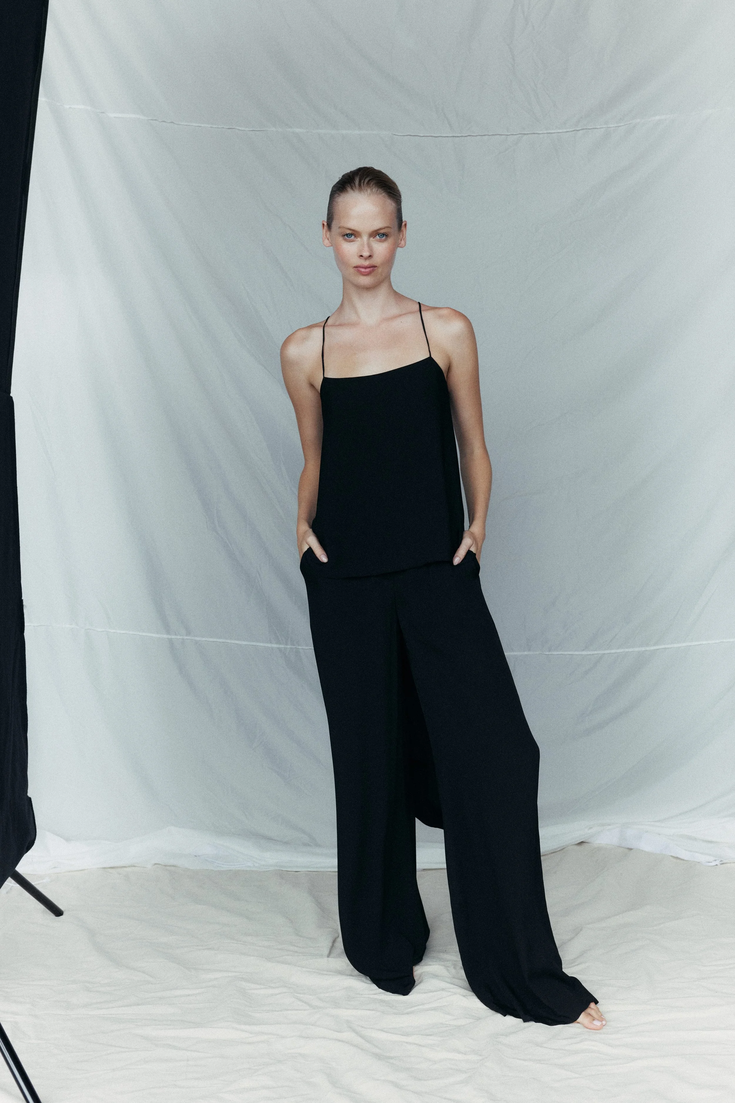 A woman in a black sleeveless top and wide-leg pants standing in front of a plain backdrop.