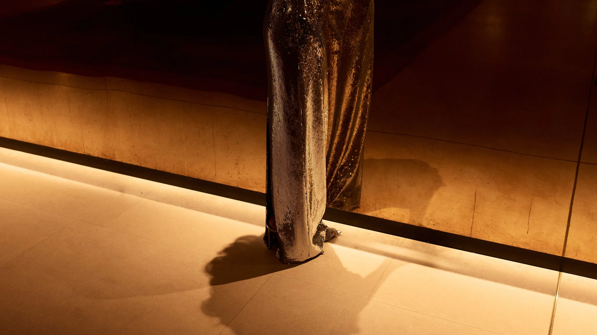Lower half of a person wearing shiny, metallic, sequined pants and metallic shoes, standing on a polished floor in front of a metallic wall.
