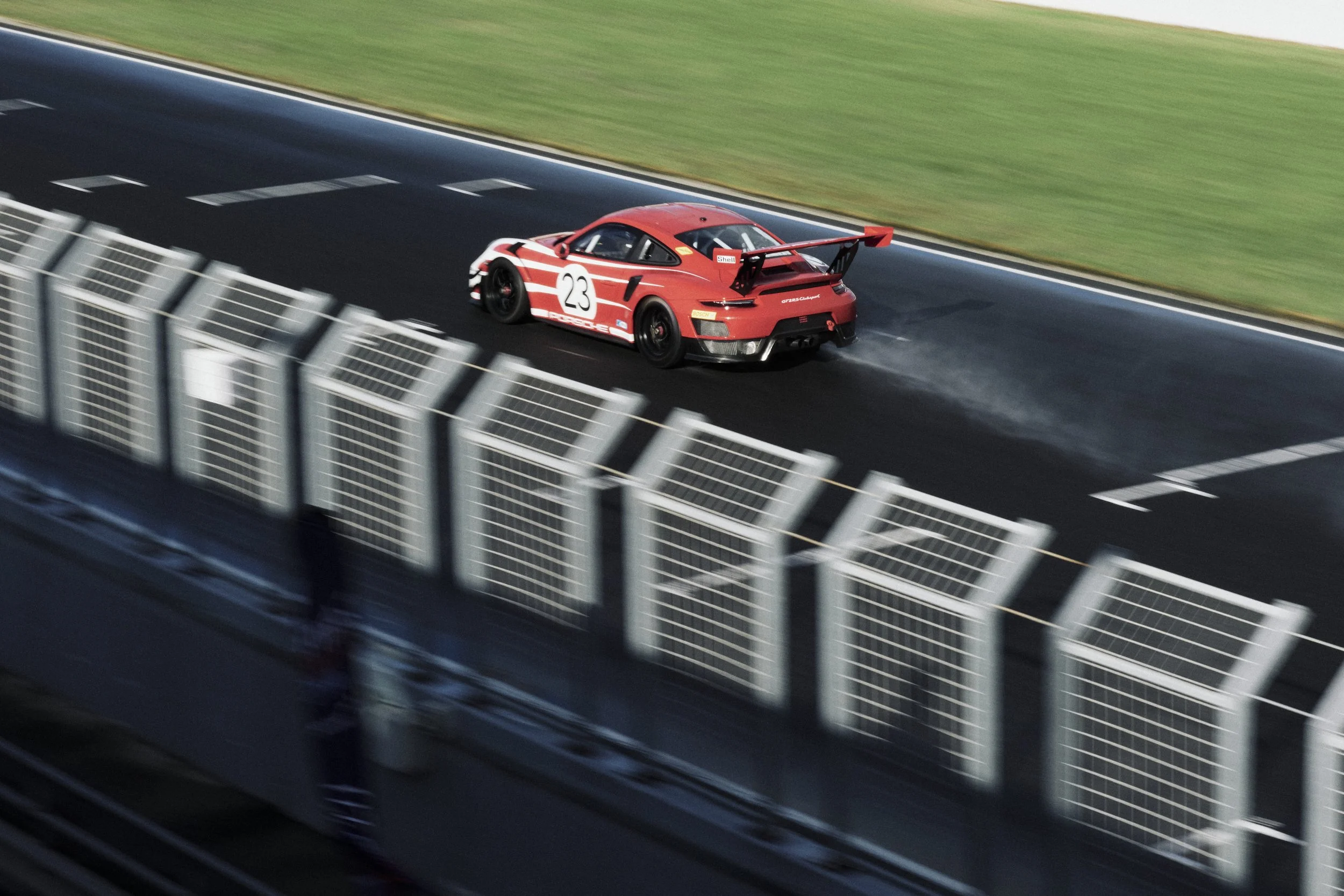 A red race car with the number 23 on its side driving on a wet race track surrounded by a safety barrier and green grass.