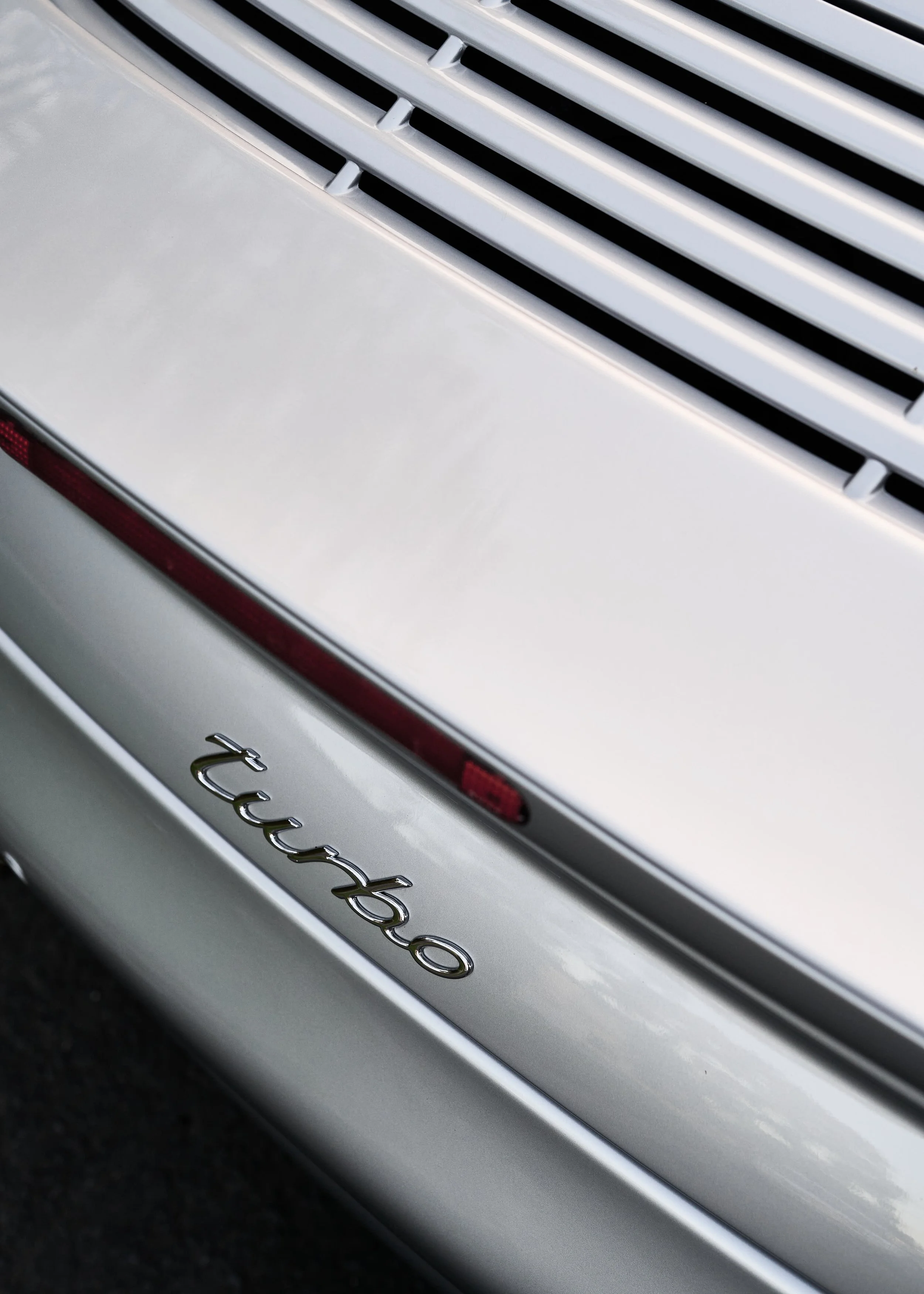 Close-up of the rear end of a silver Porsche Turbo, showing the Turbo badge and part of the taillight with horizontal vents above.