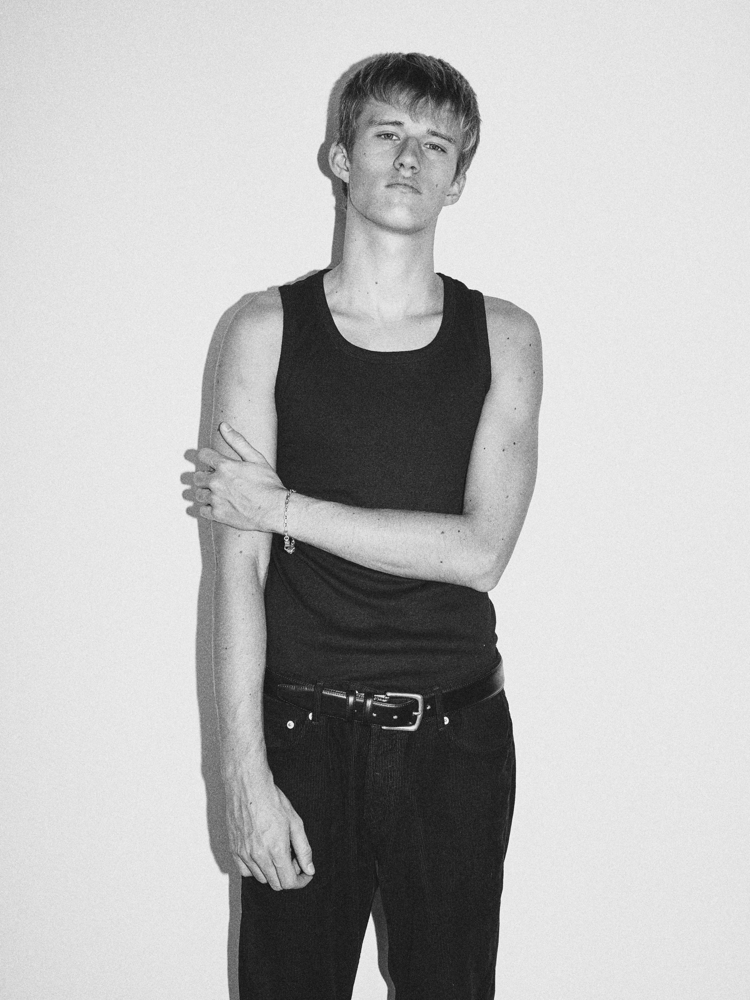 Black and white portrait of a young person with short hair, wearing a sleeveless black shirt and dark jeans, standing against a plain background.