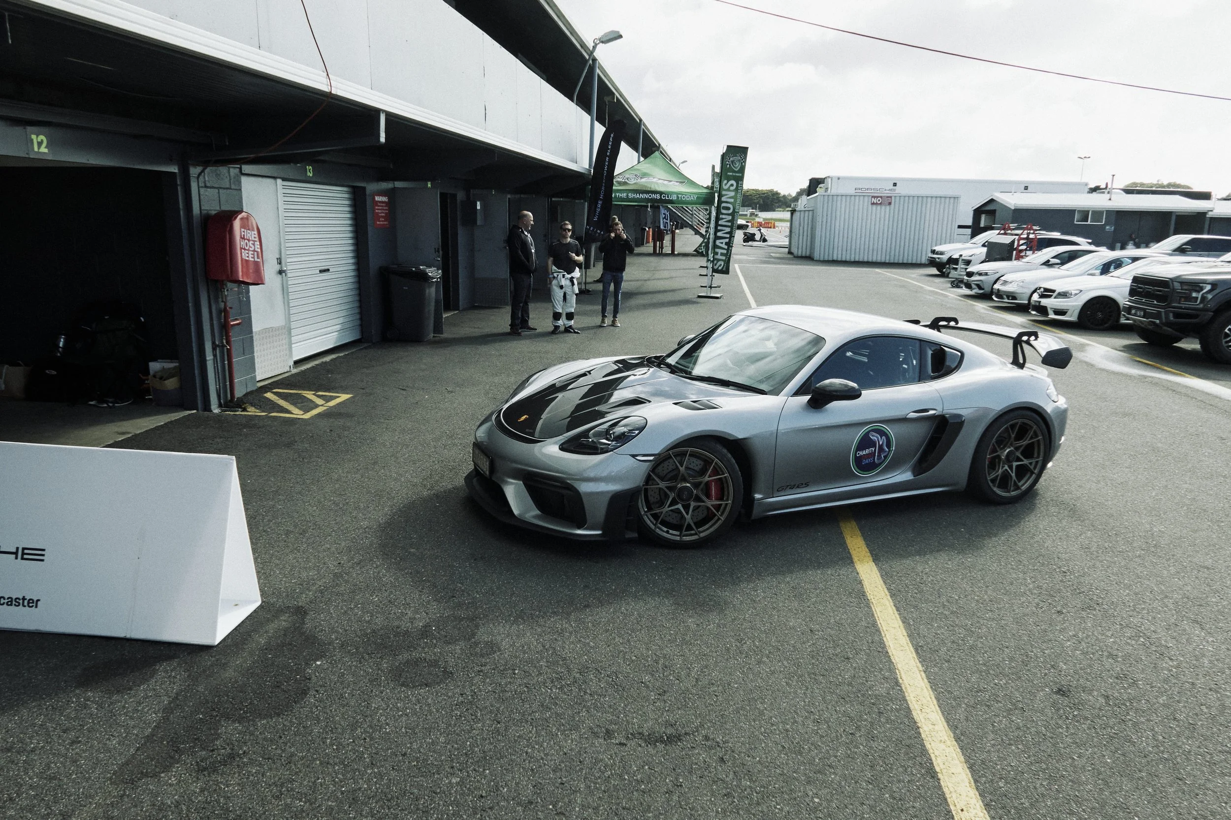 A silver racing sports car with black and carbon fiber accents parked in a paddock area at a racetrack, with a group of three people standing nearby and several other cars parked in the background.