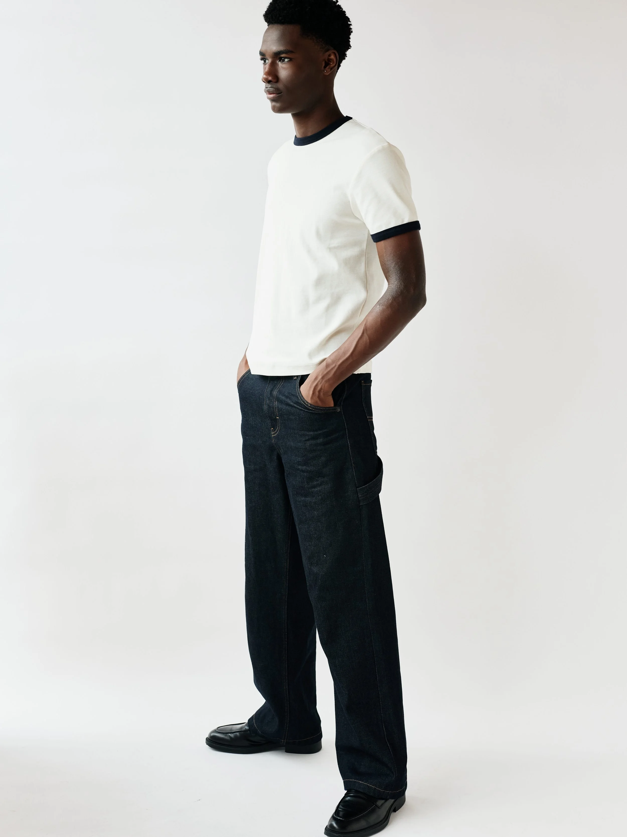 Young man standing in profile, wearing a white t-shirt with black trim and dark jeans, against a plain white background.
