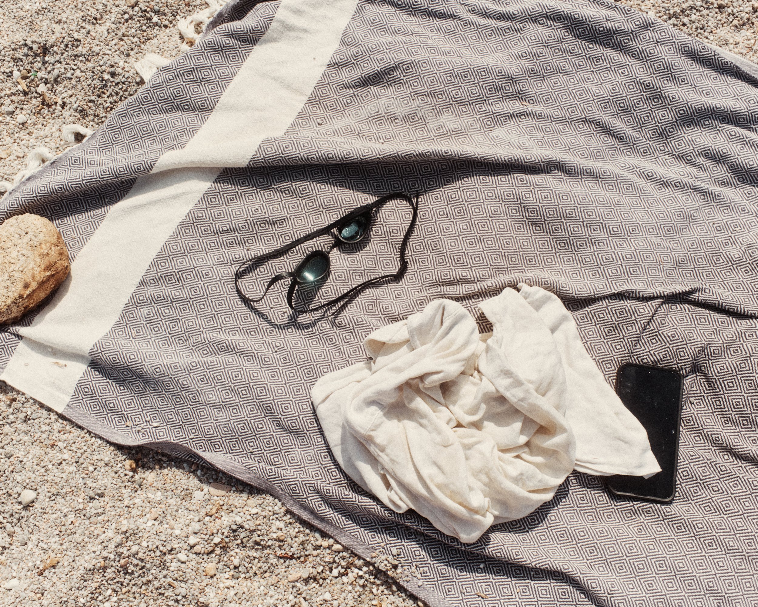 A patterned towel on sandy ground with a rock, a pair of sunglasses with blue lenses, a white cloth, and a black smartphone.