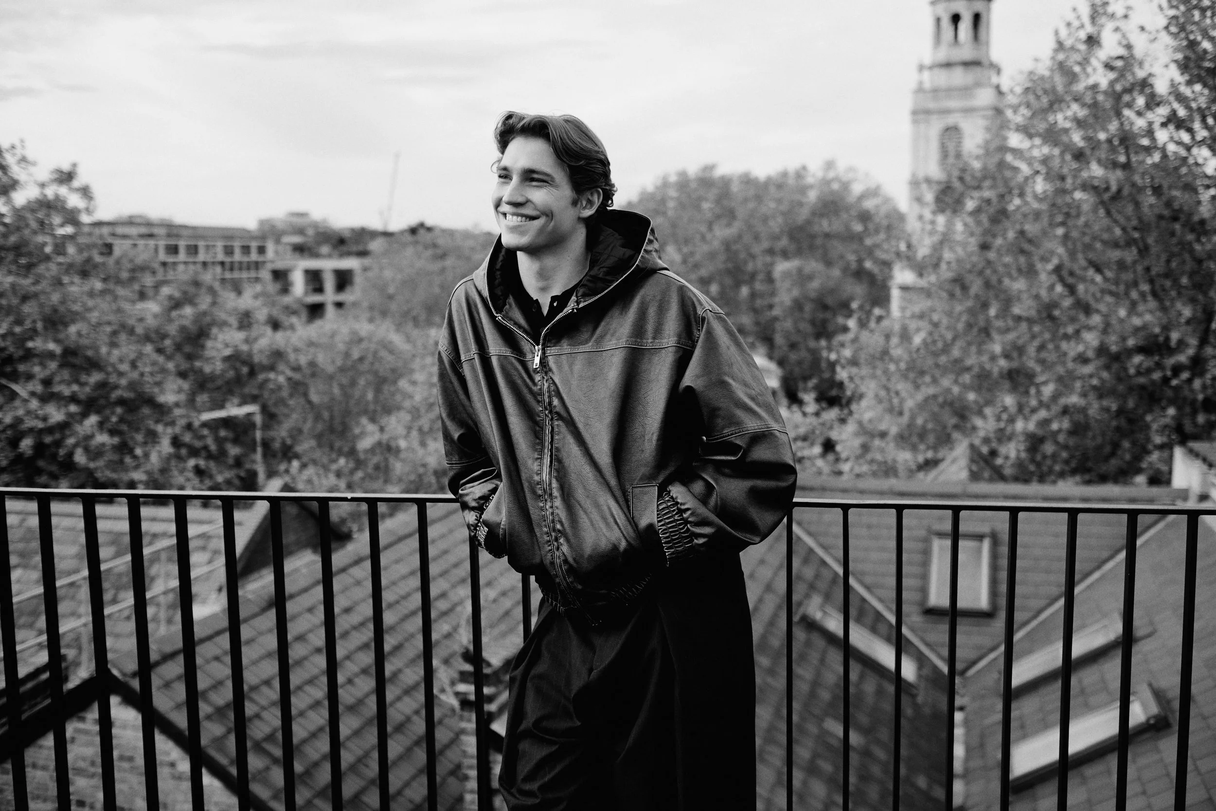 Young man in a leather jacket smiling and leaning on a balcony railing with trees and a building tower in the background, black and white photo.