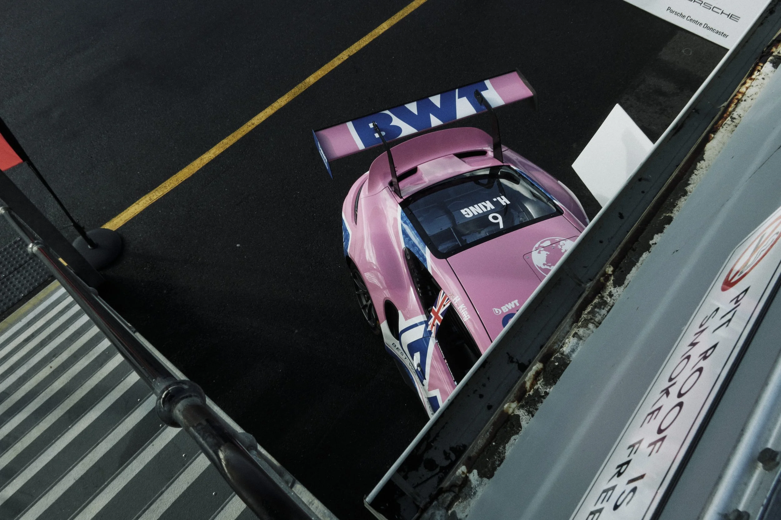 A pink racing car with a large rear wing parked in the pit lane at a racetrack, viewed from above.