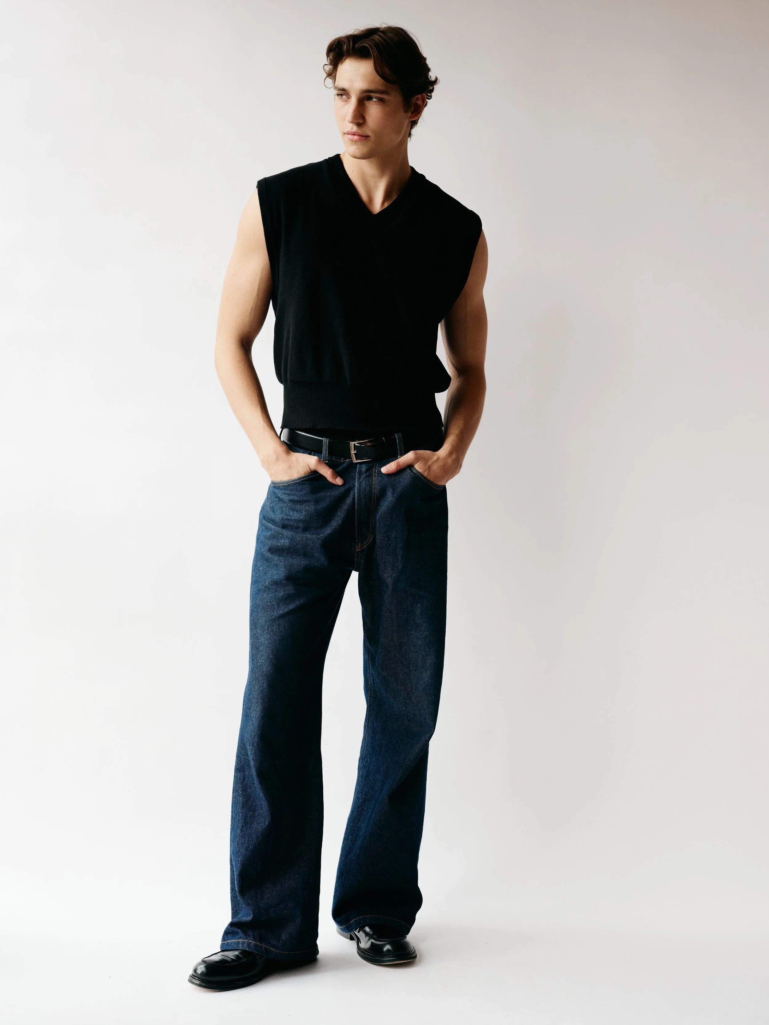 Young man wearing a sleeveless black sweater, blue jeans, and black shoes, standing with hands in pockets against a plain light background.