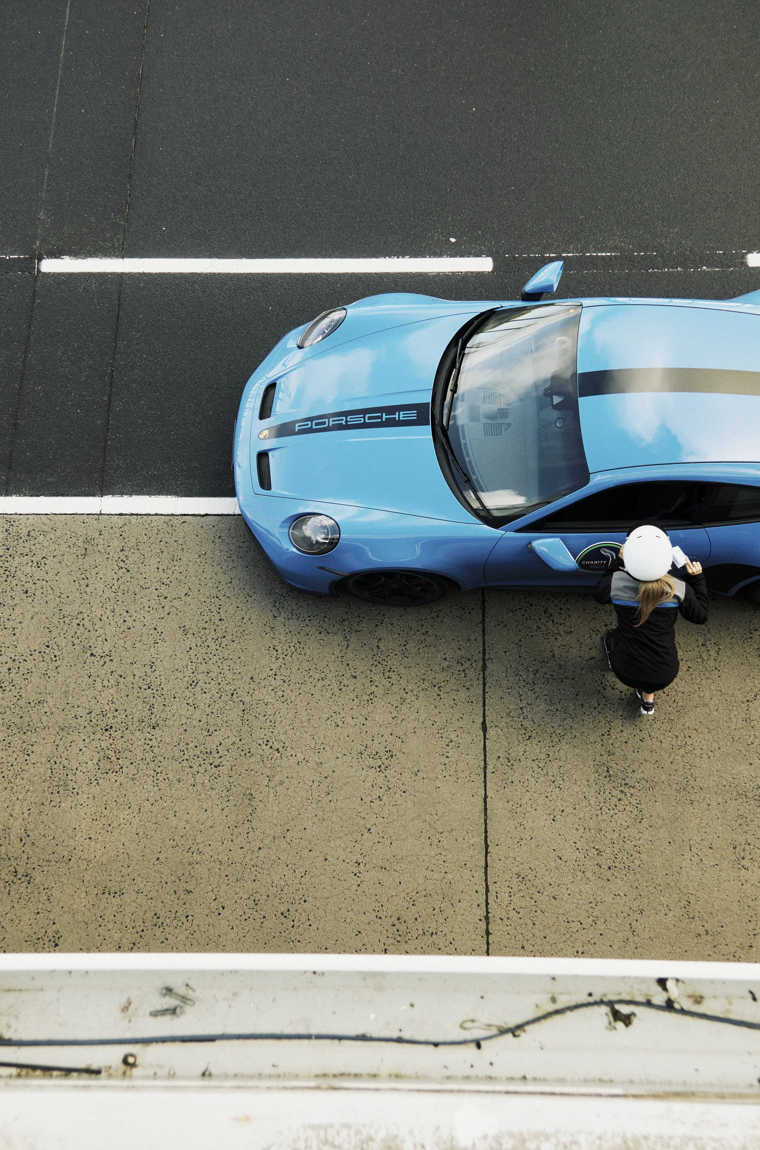 Bird's eye view of a blue Porsche car parked on a parking lot, with a woman crouching next to the car, holding a helmet.